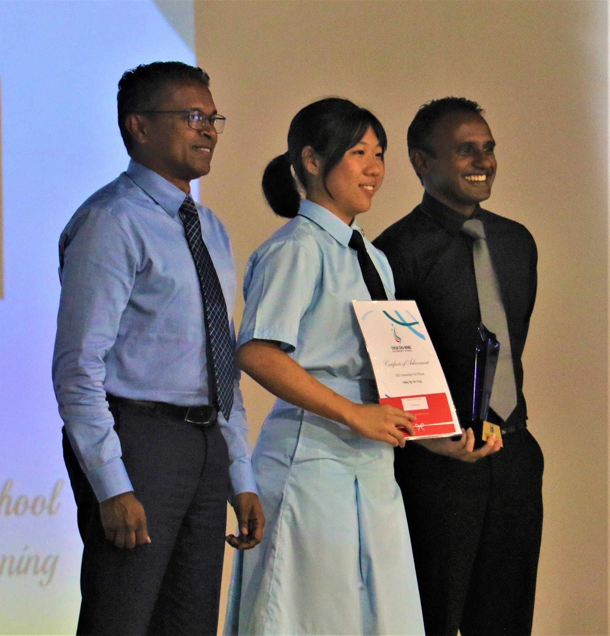 A smiling student in a light blue uniform holds a certificate on stage, flanked by two men in formal attire.