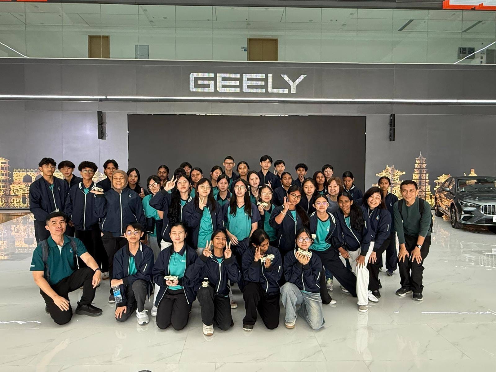 A large group of people in matching teal and navy uniforms pose together inside a Geely automotive showroom.
