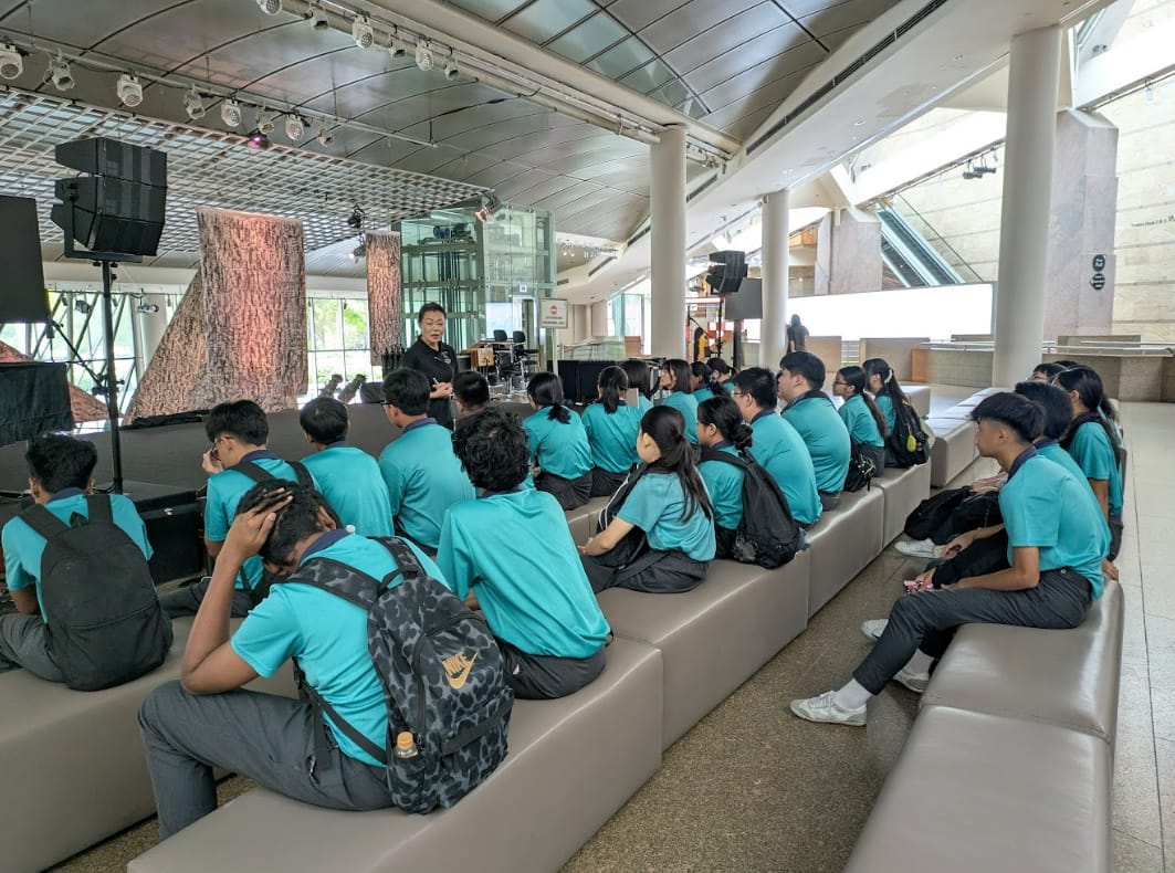 Students in matching teal shirts sit on benches facing a presenter standing near a large tree bark display indoors.