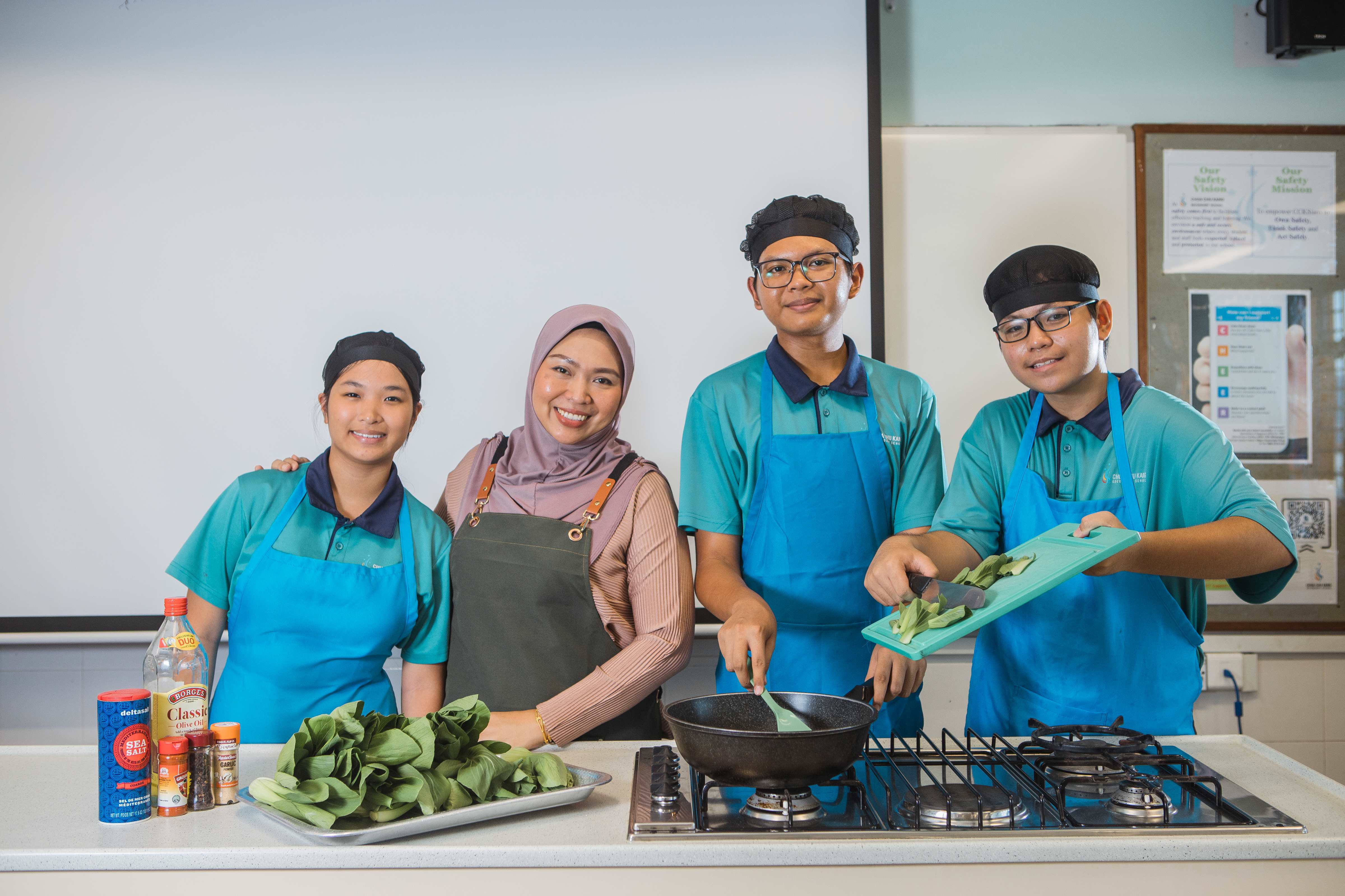 Four people smile while cooking green vegetables in a pan at a kitchen counter with spices nearby.