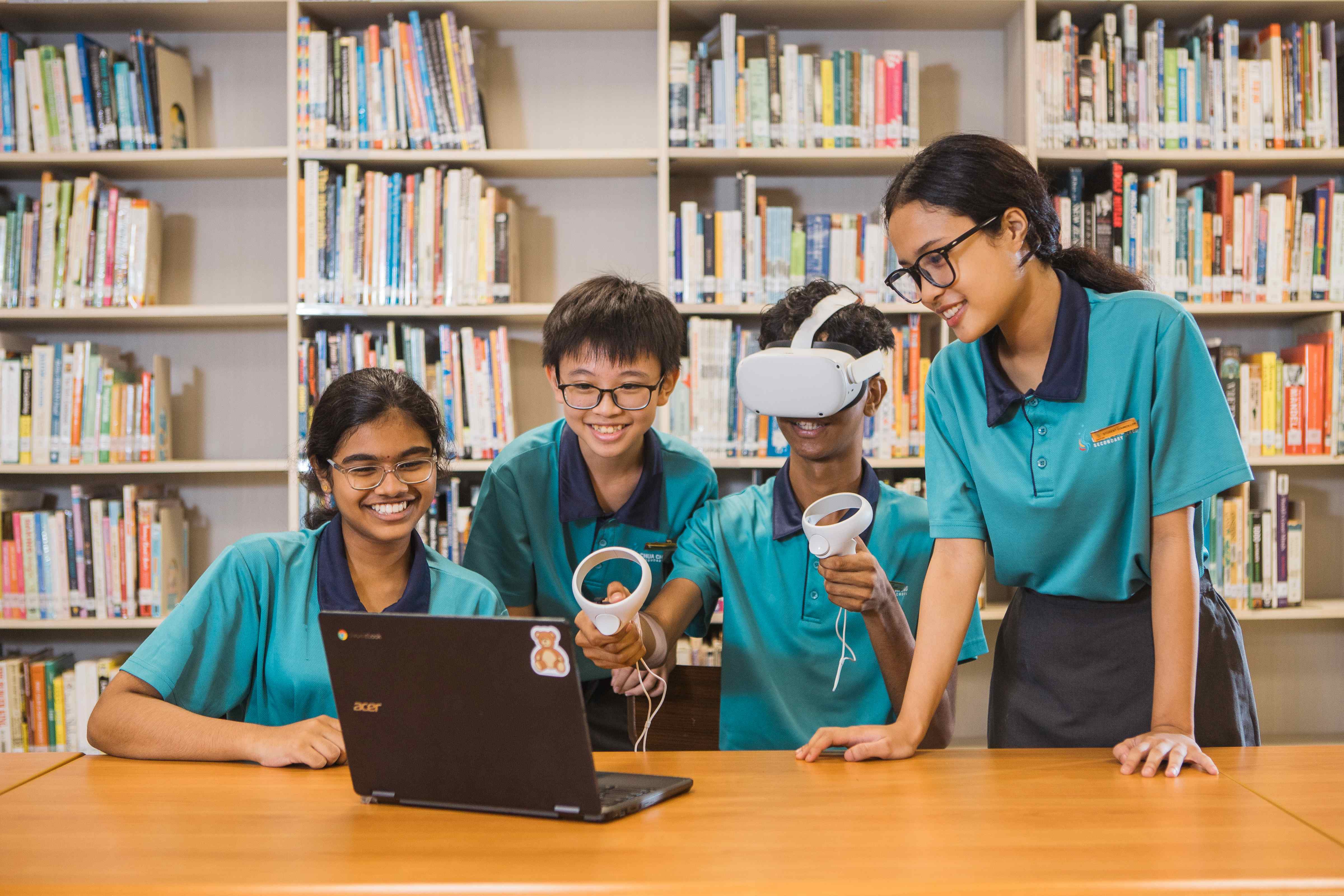 Four students in teal uniforms use a VR headset and laptop in a library with bookshelves behind them.