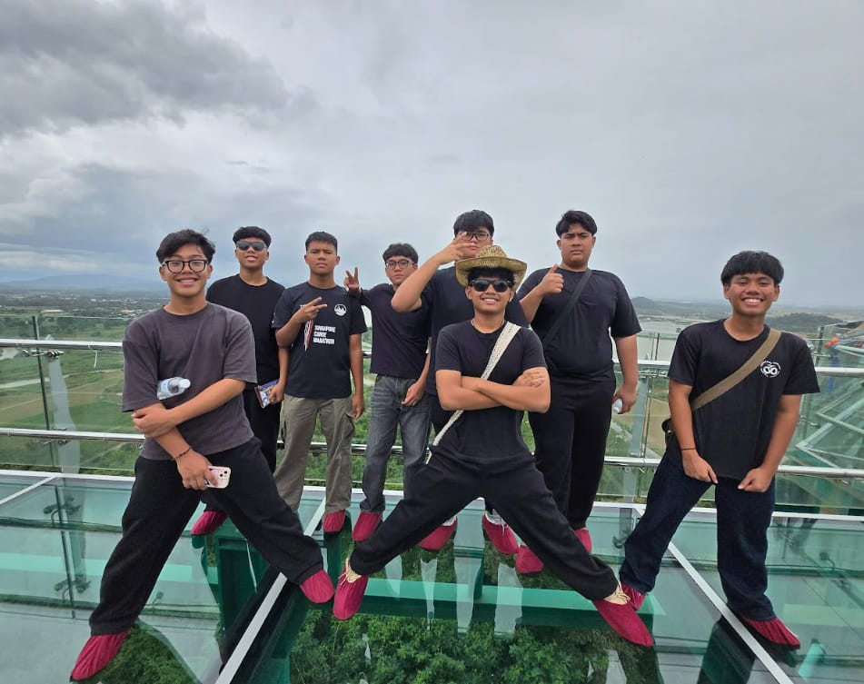 Eight young men in black outfits pose on a glass floor observation deck with a scenic landscape below.