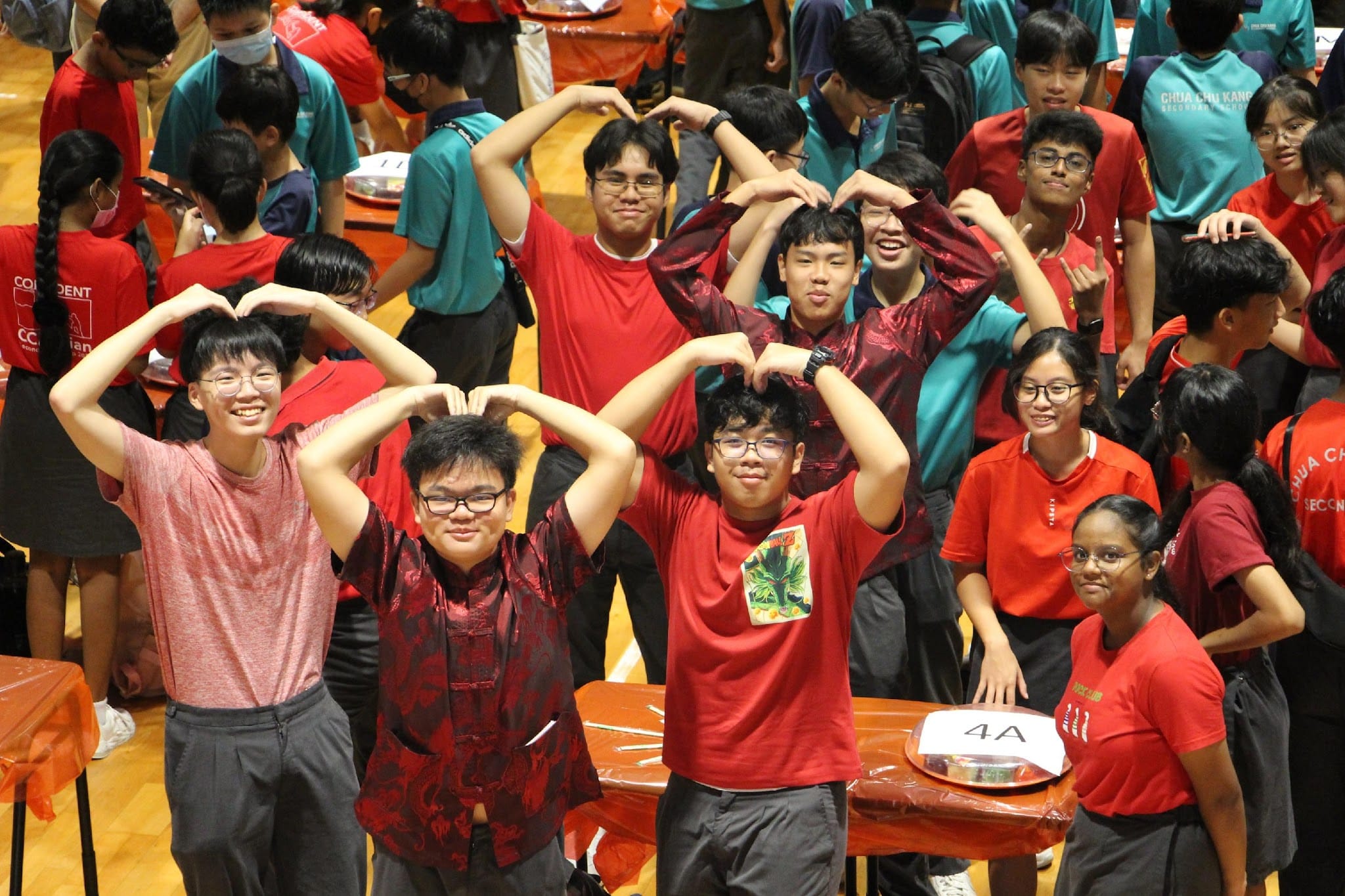 Group of students in red and teal shirts forming heart shapes with their hands at a school event, table labeled 4A visible.
