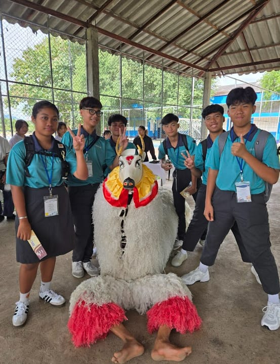 Six students in teal uniforms pose with a decorative bird costume under an outdoor shelter.