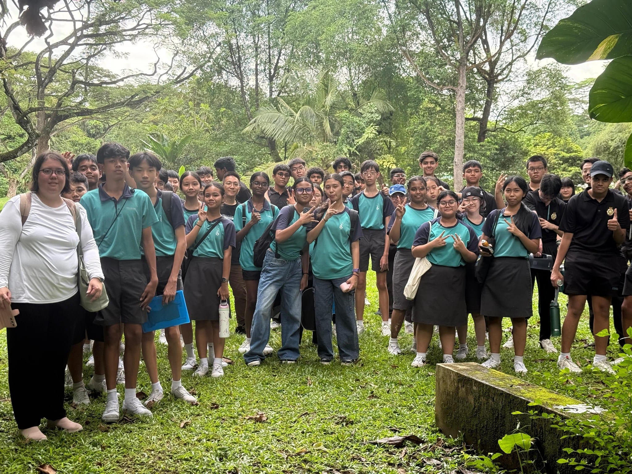 A large group of students in teal uniforms poses outdoors on grass surrounded by lush green trees.