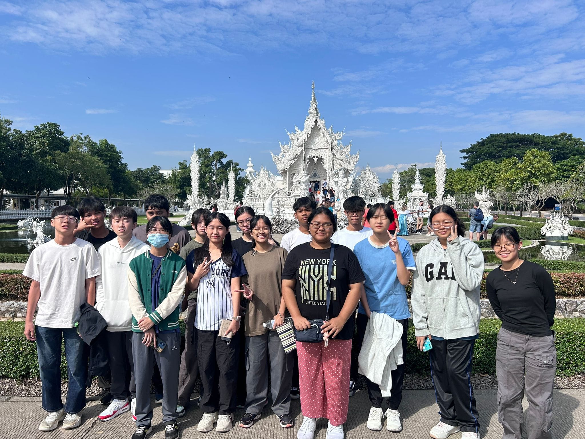 A group of teenagers posing in front of the ornate white Wat Rong Khun temple in Thailand under a blue sky.
