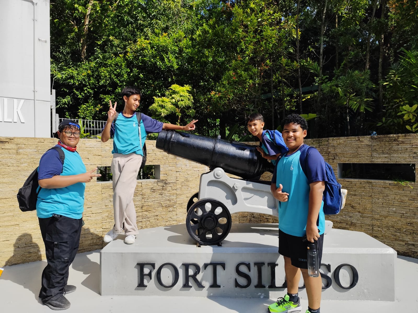 Four boys in teal shirts pose with a black cannon at Fort Siloso sign, surrounded by green trees.