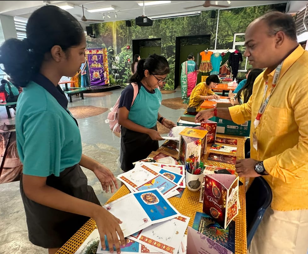 Two students in teal uniforms browse Diwali cards and decorations at a table with a man in a yellow kurta.