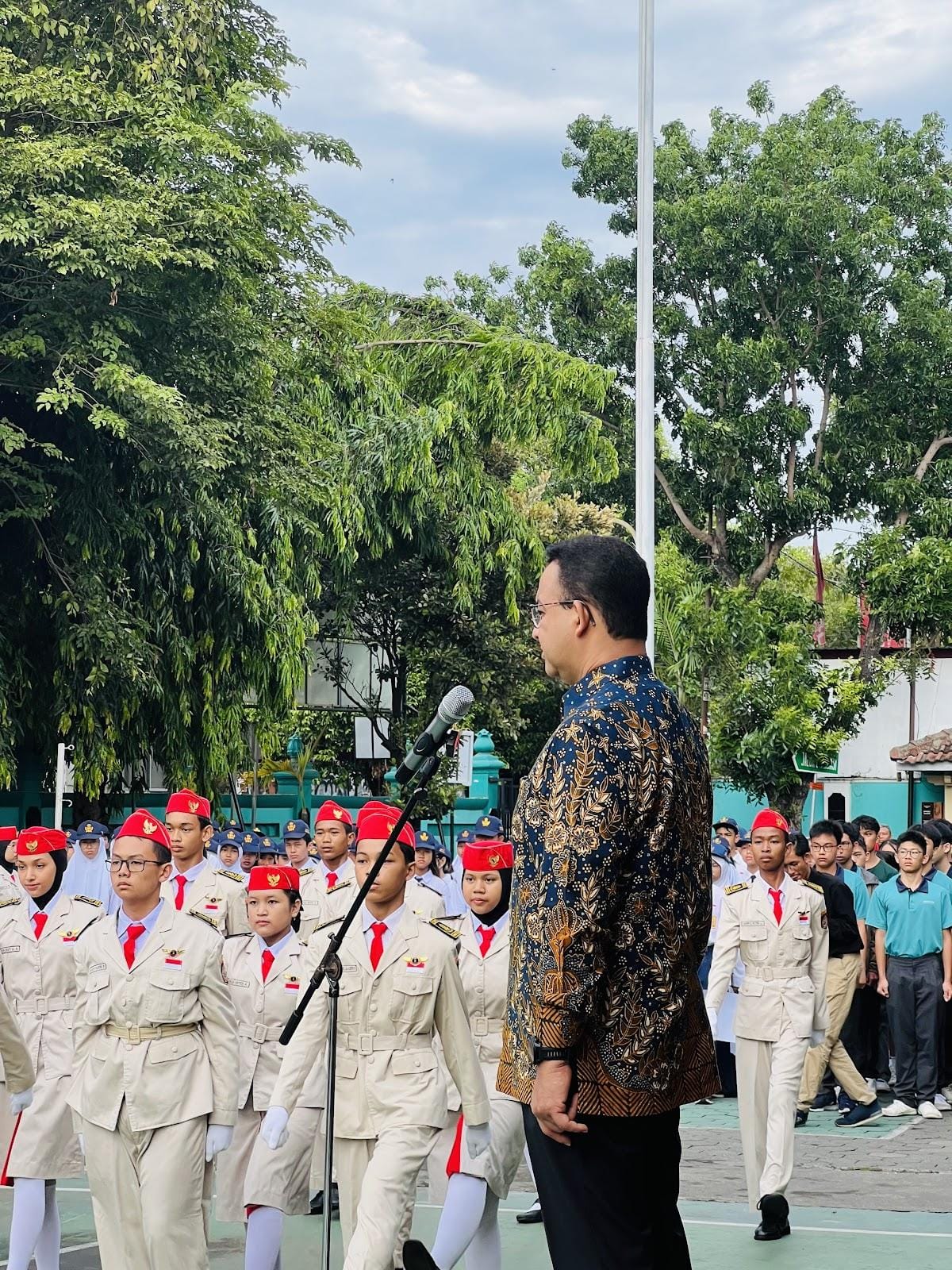 A man in a batik shirt speaks at a microphone before rows of youth in white uniforms and red caps outdoors.