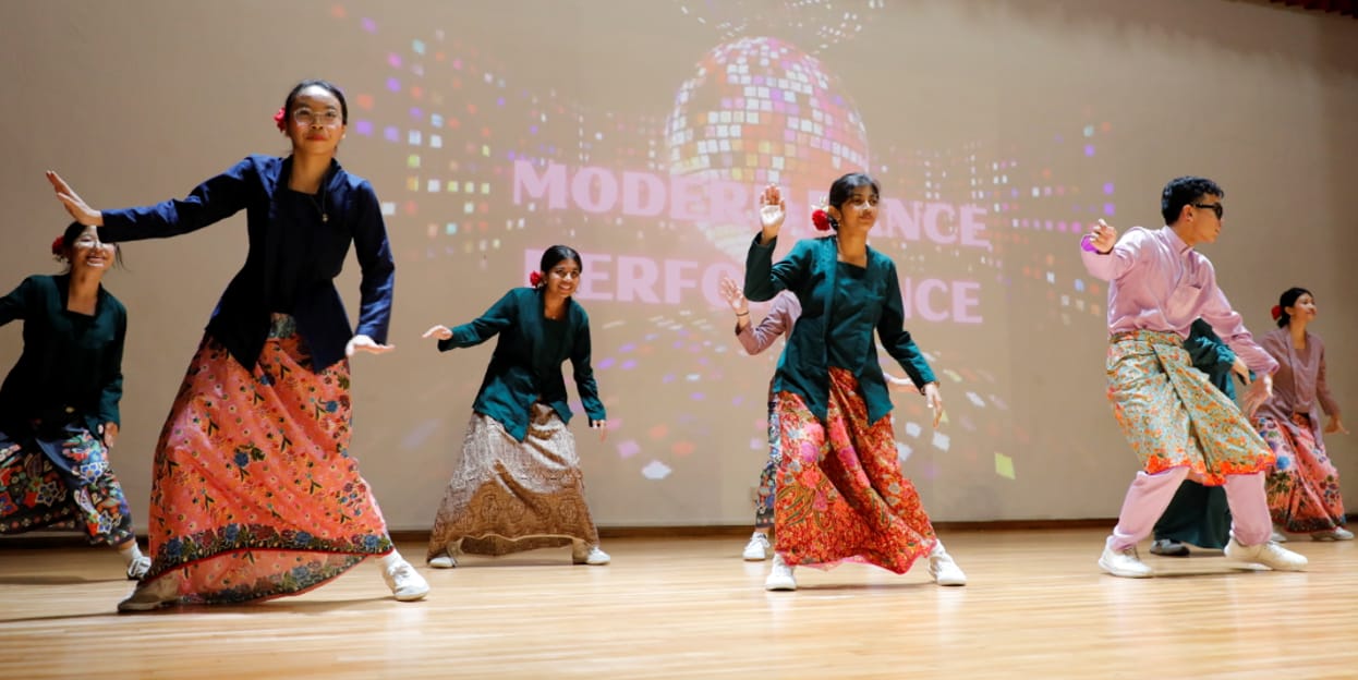 Six dancers in colorful skirts and jackets perform on stage before a projected "Modern Dance Performance" backdrop.