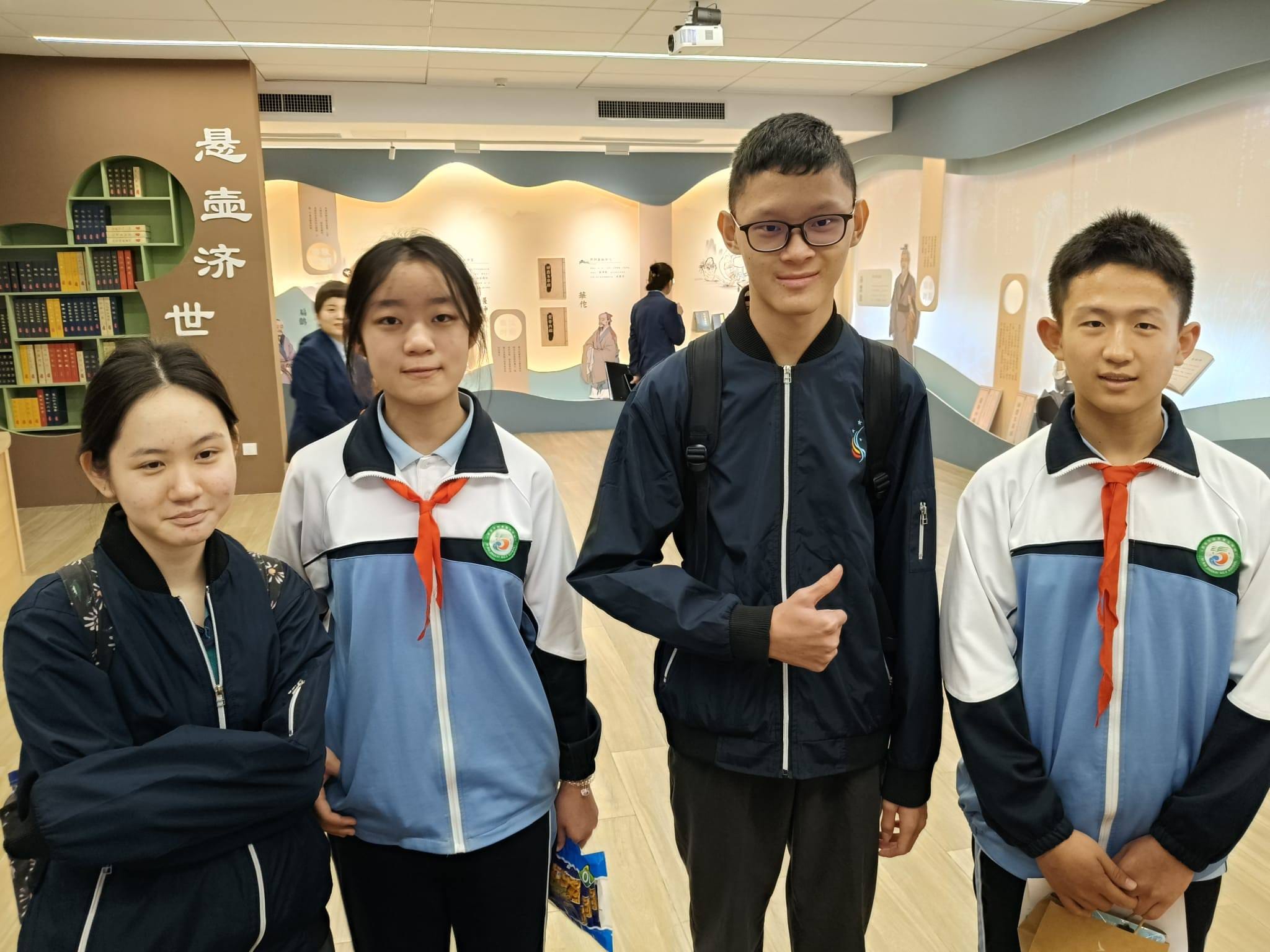 Four young people pose in a Chinese cultural exhibition hall with Chinese characters and bookshelves visible behind them.