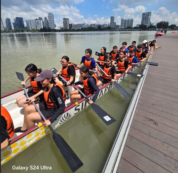A group of young paddlers in orange life vests sit in a long dragon boat on a river near a city skyline.