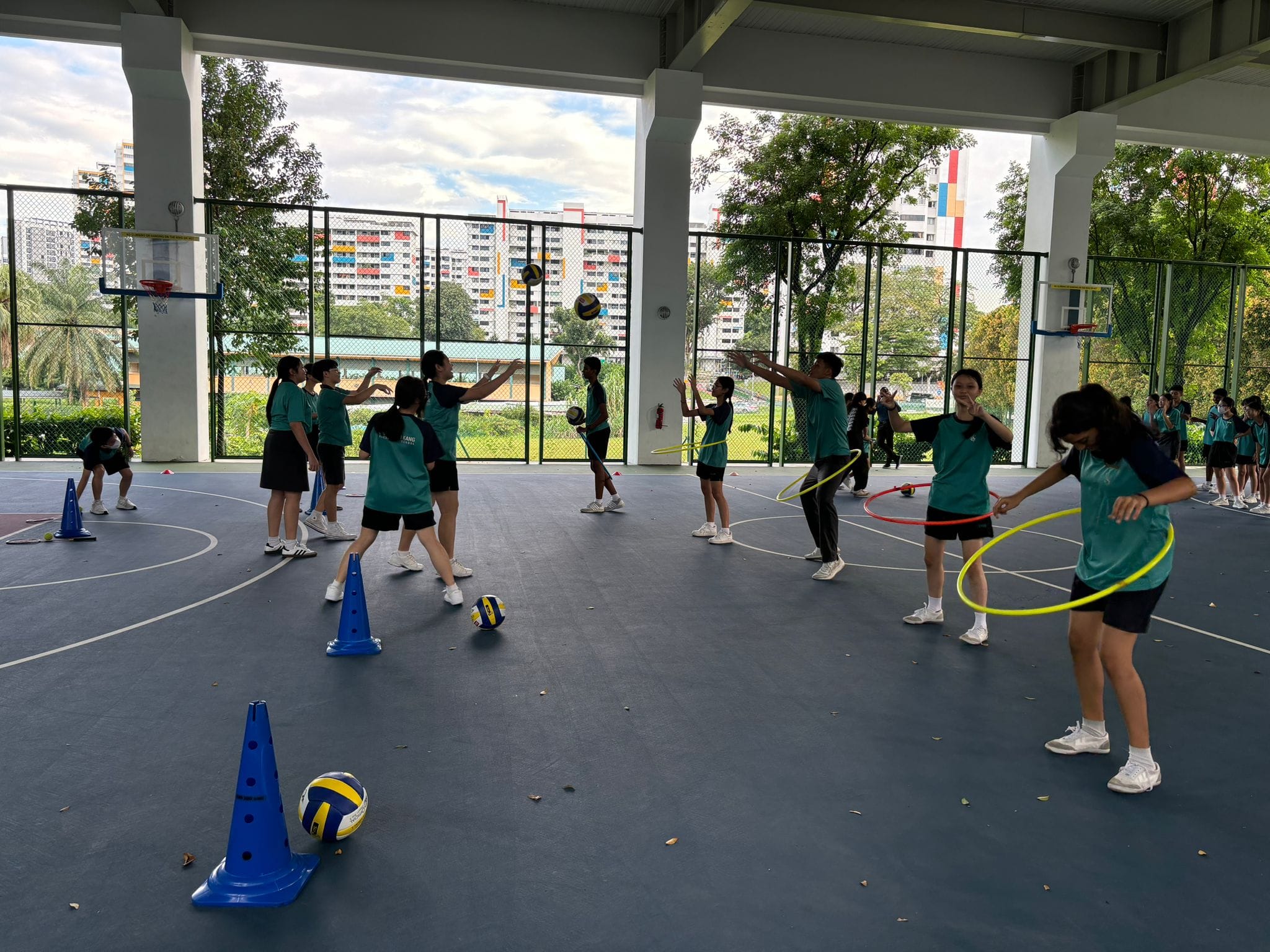 Students in teal shirts practice on a covered court with volleyballs, hula hoops, and blue cones.