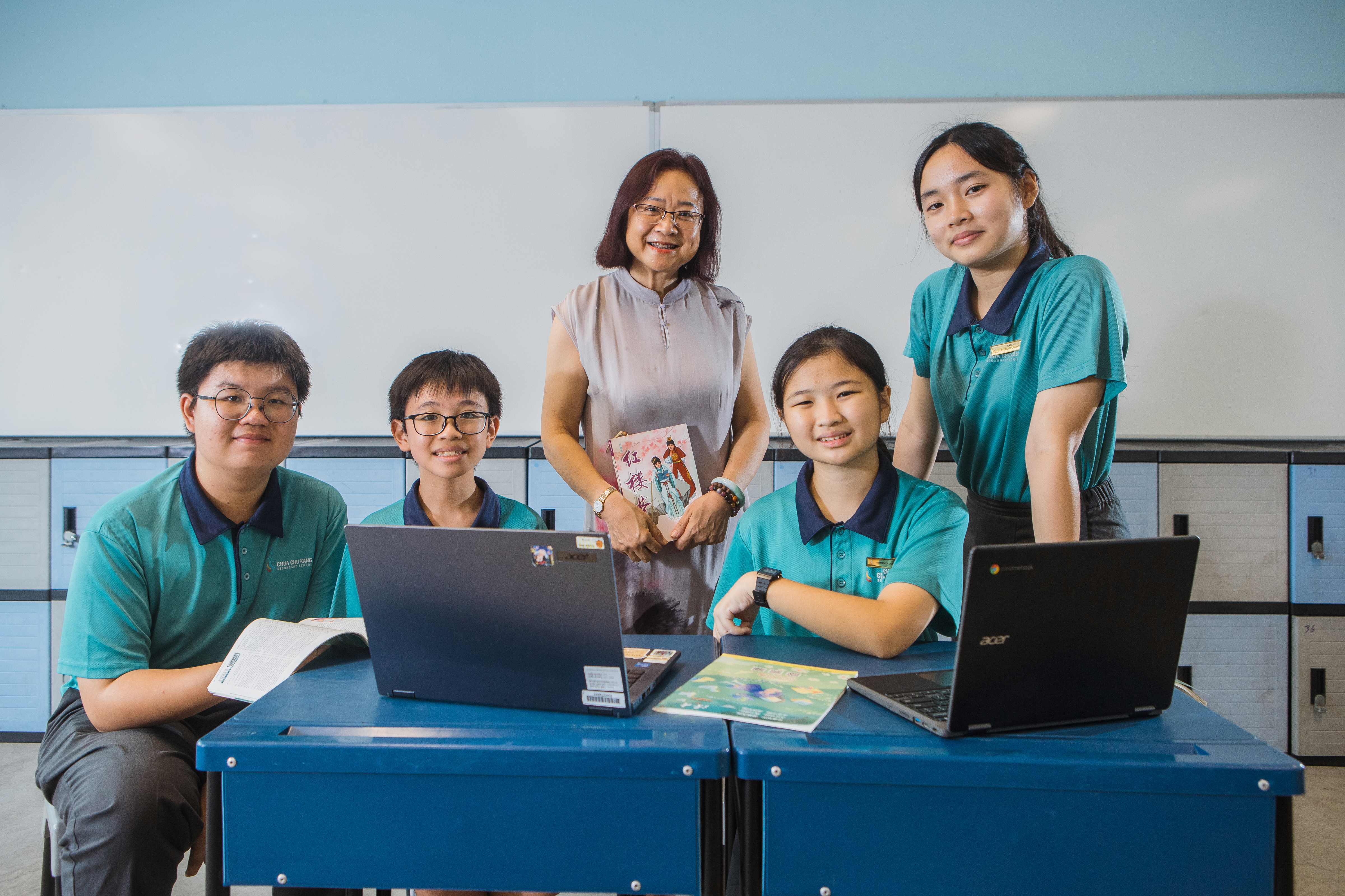 A teacher holding a book stands with four students in teal uniforms around a blue desk with laptops in a classroom.