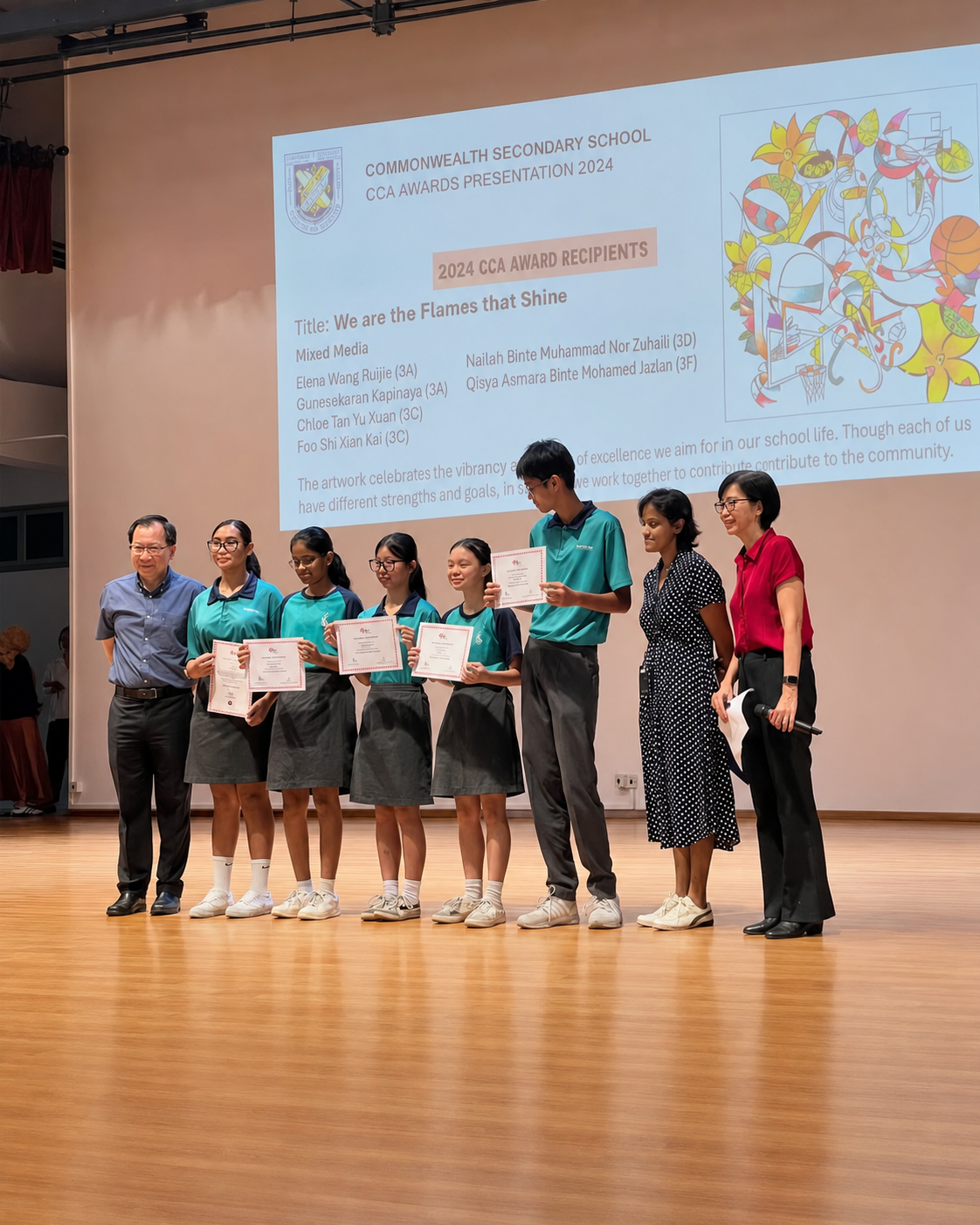 Students in teal uniforms hold certificates on a stage in front of a projected slide titled "We are the Flames that Shine."