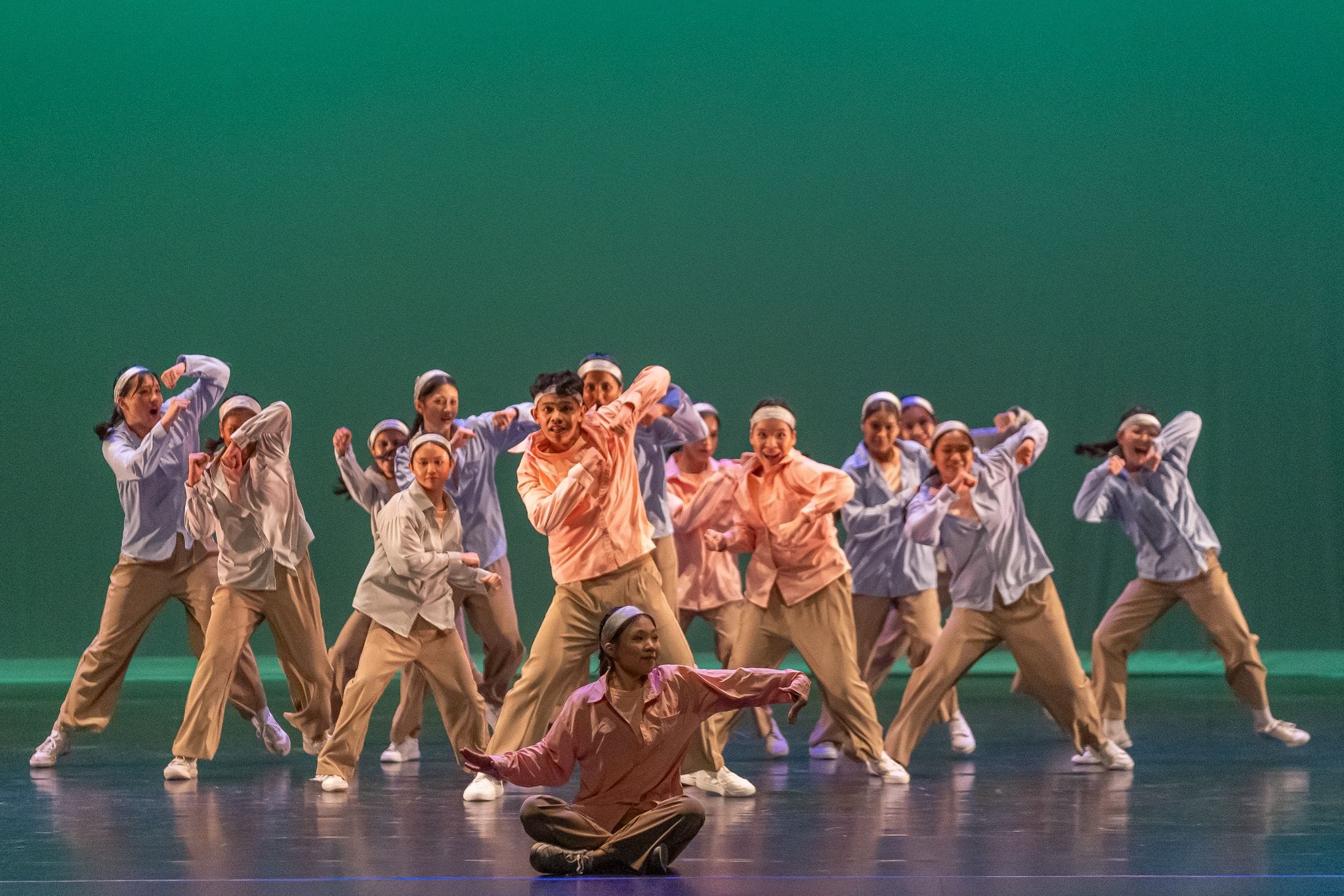 A large group of dancers in blue and pink shirts perform on a stage with a green backdrop, one seated in front.