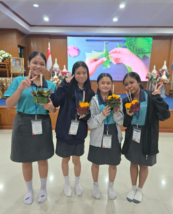 Four girls in school uniforms with lanyards pose with peace signs, each holding a decorative krathong float indoors.