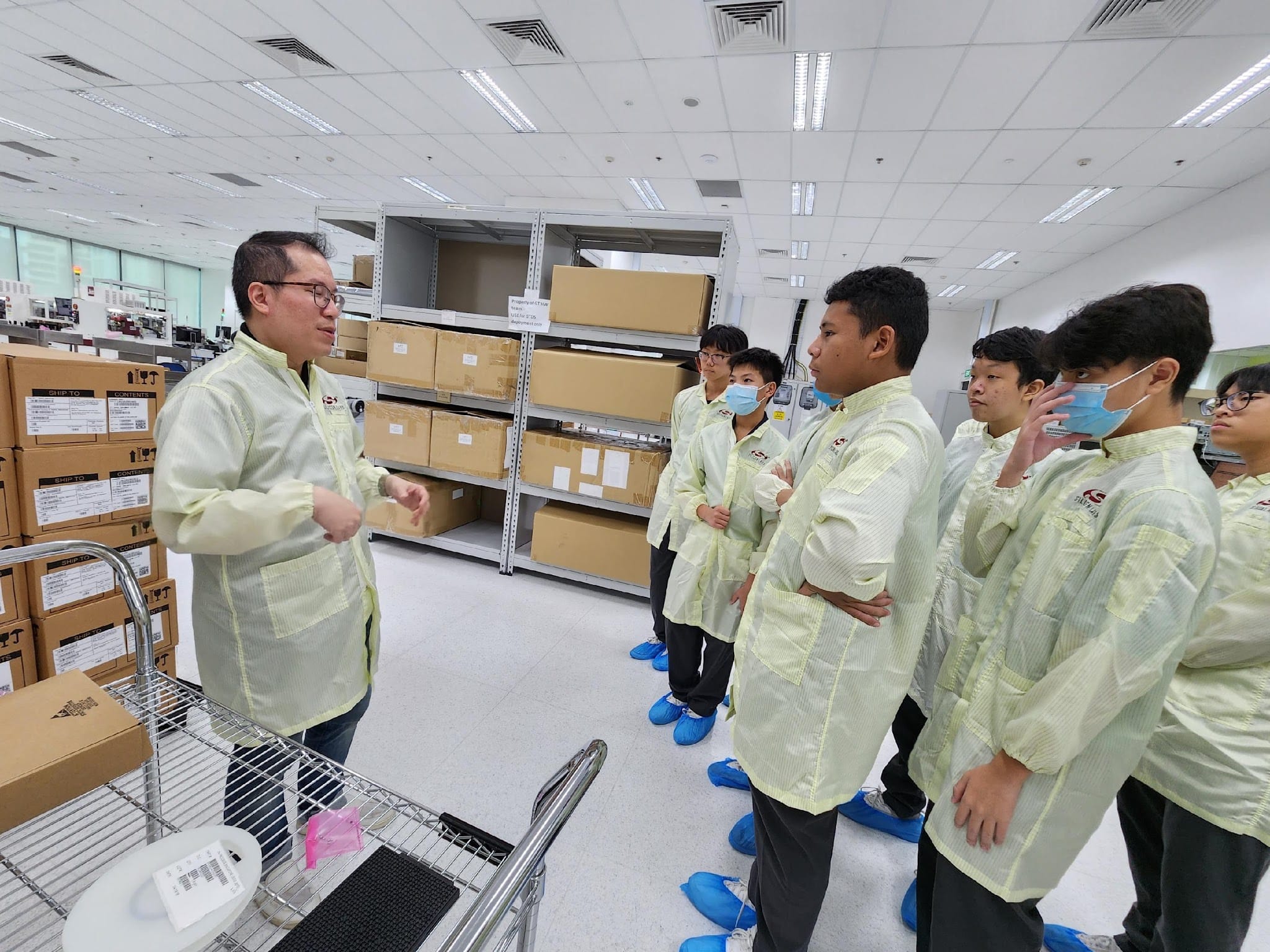 A man in a yellow lab coat speaks to a group of students wearing matching coats and blue shoe covers in a warehouse facility.