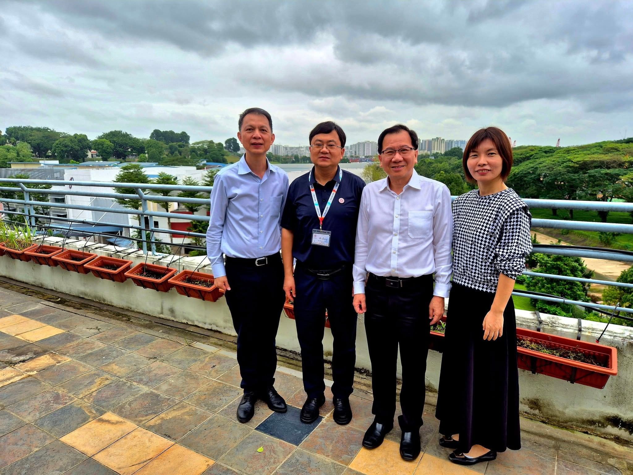 Four people stand on an outdoor rooftop terrace with potted plants, trees, and a cloudy sky in the background.