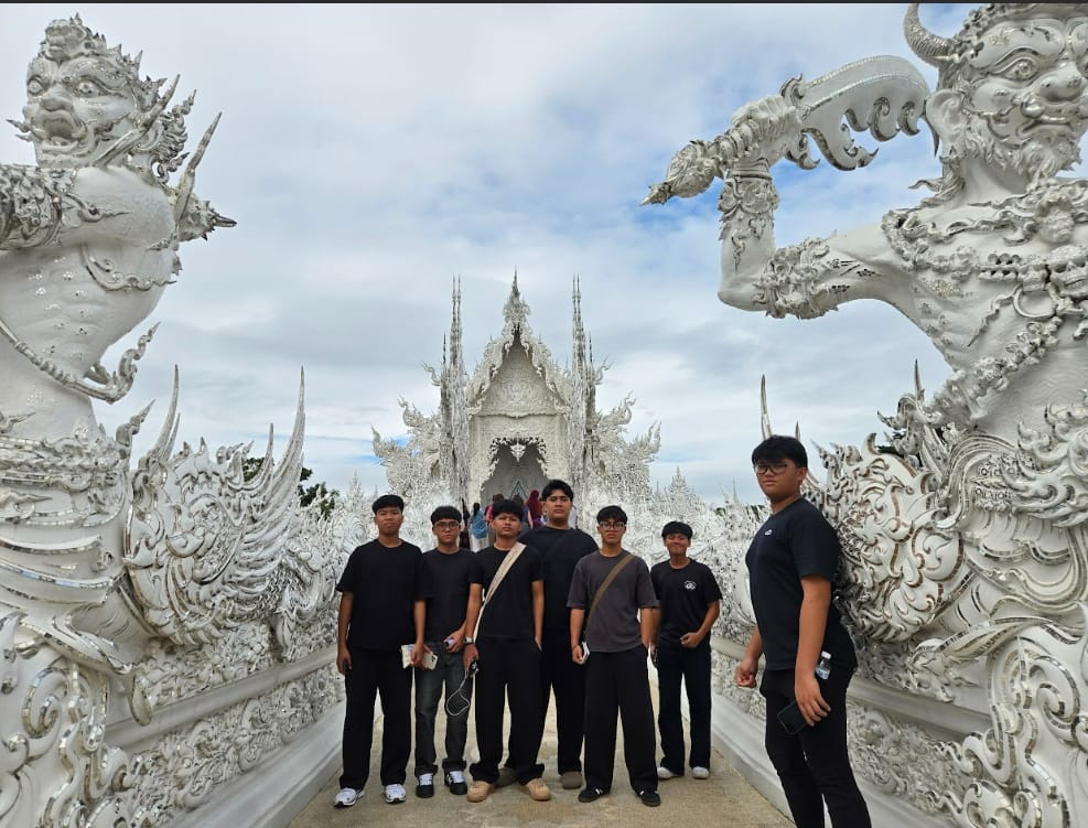 Six young people in dark clothing pose before the ornate white Wat Rong Khun temple in Thailand.