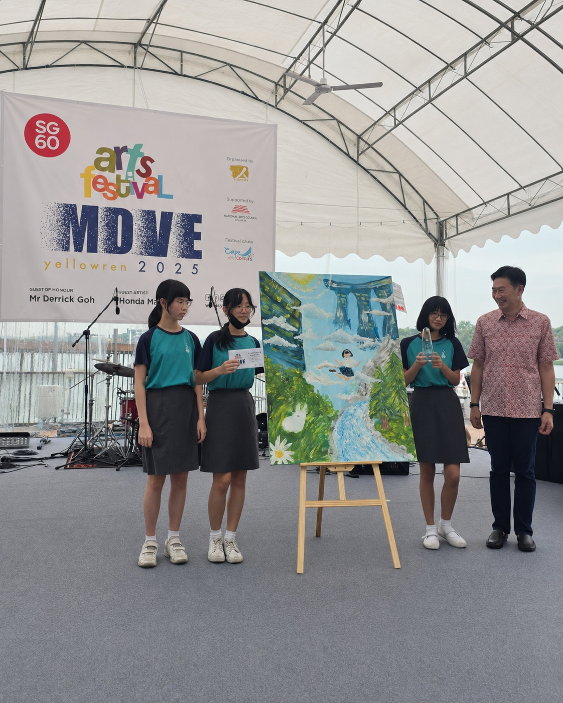 Three students in teal uniforms stand beside a colorful painting on an easel at the SG60 Arts Festival MOVE event.