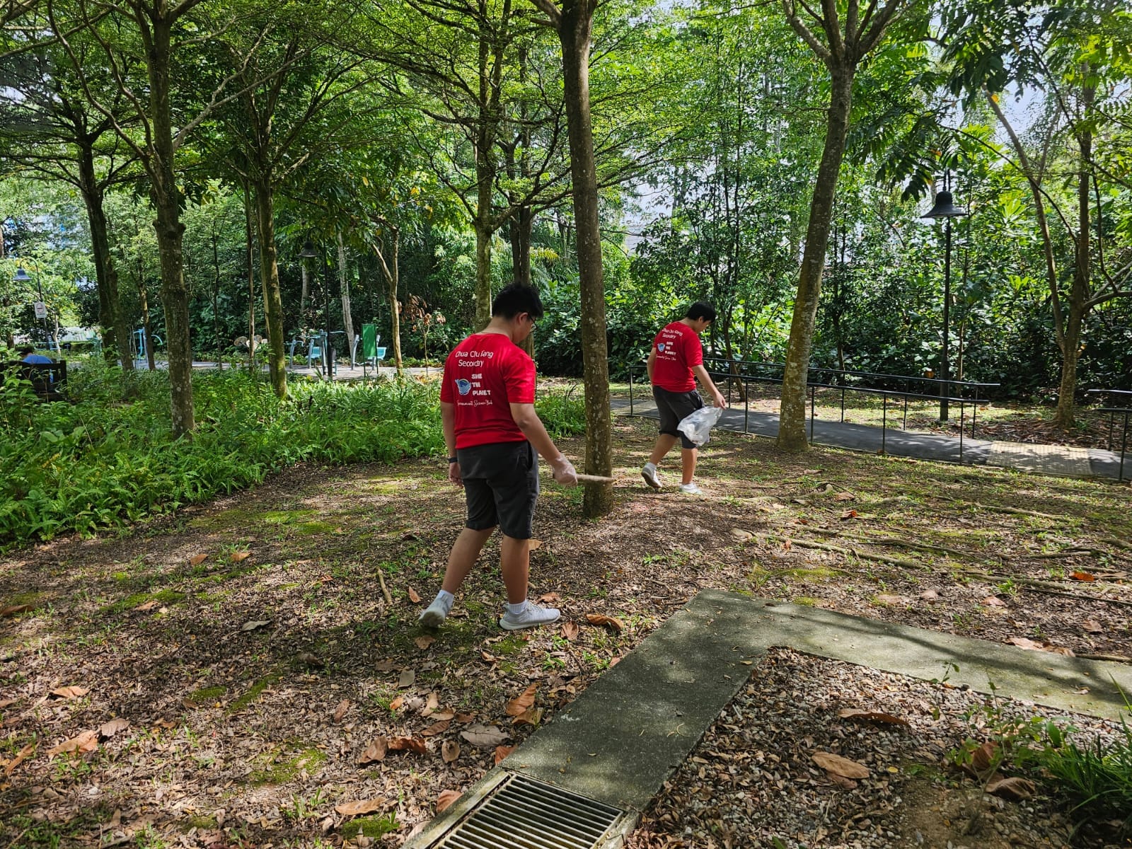 Two boys in red shirts pick up litter with a stick and bag in a tree-lined park.