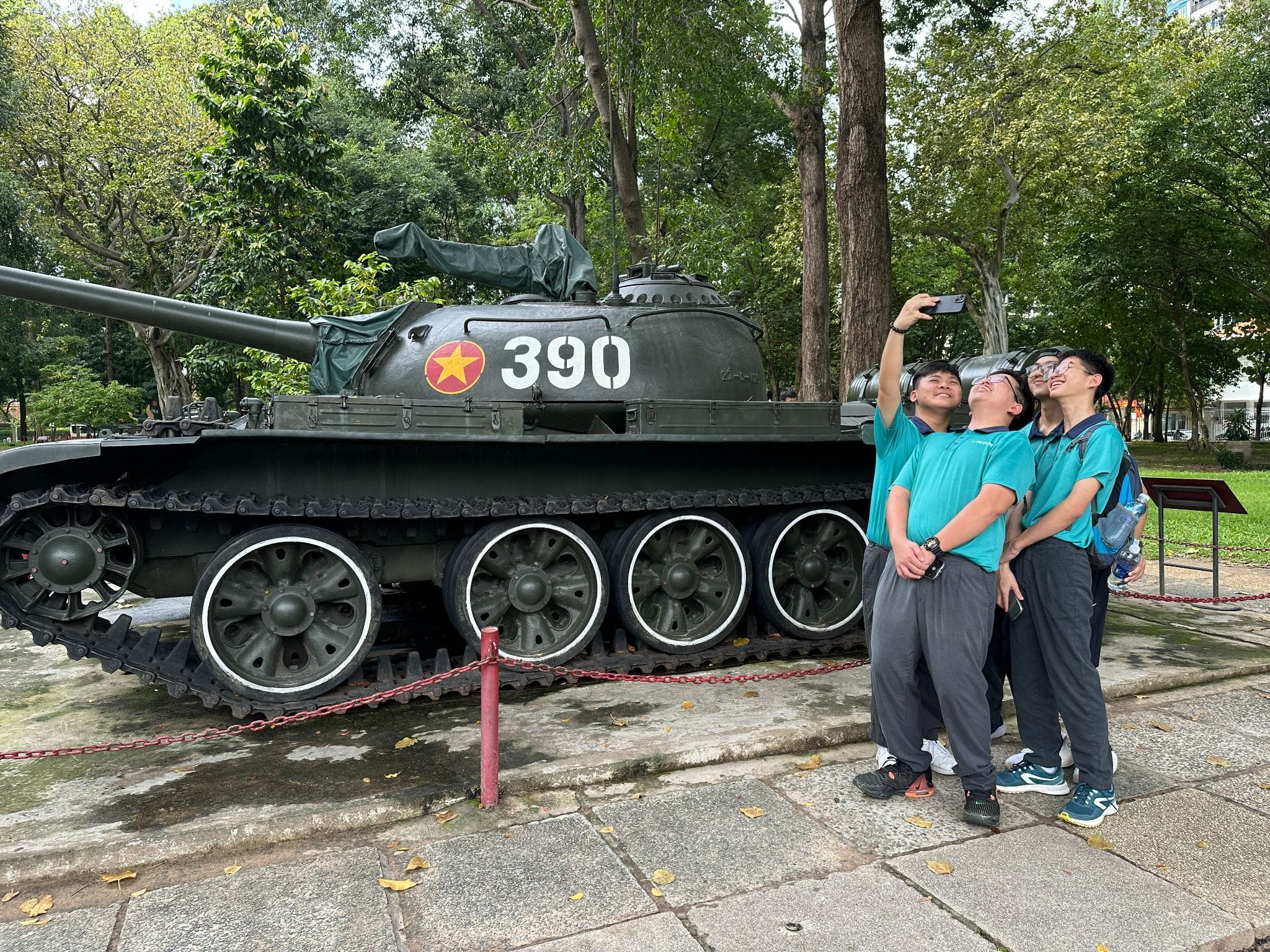Three young people in teal shirts take a selfie beside a dark green military tank numbered 390 with a red star emblem.