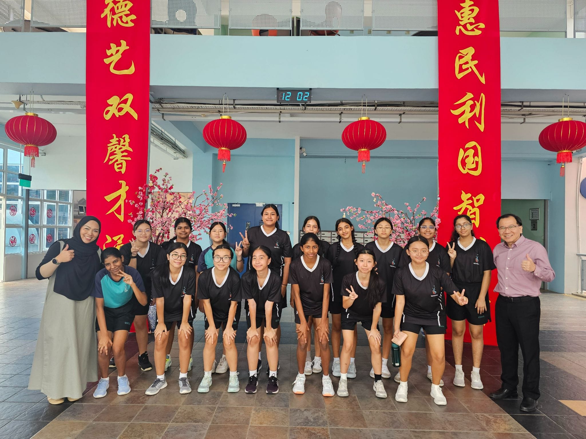 A group of students in black sports uniforms pose indoors with red Chinese New Year banners and lanterns.