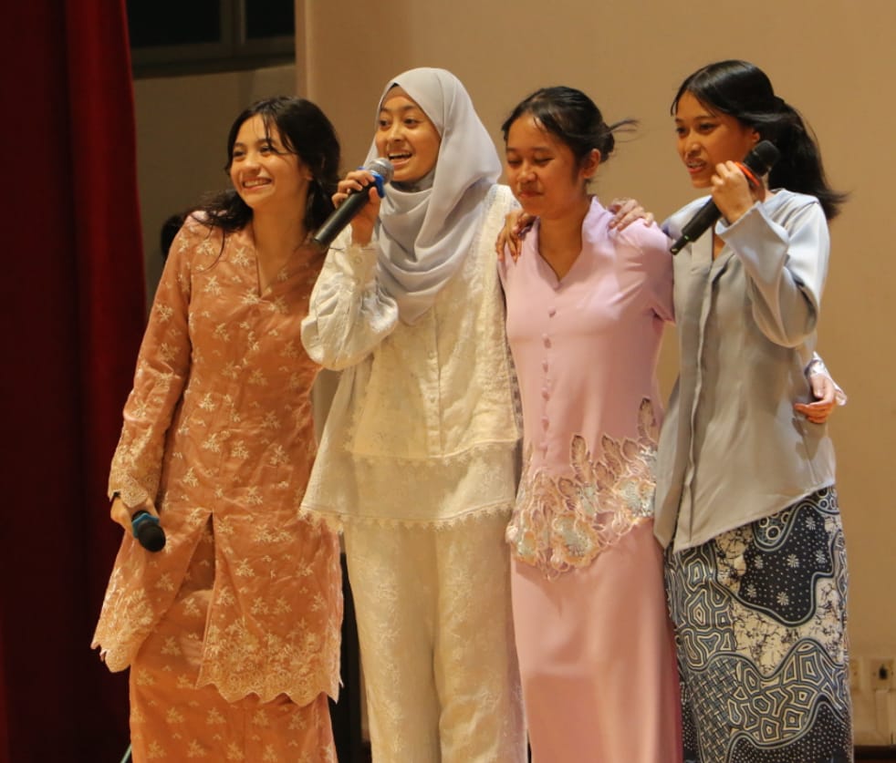 Four young women in traditional Southeast Asian attire stand together on stage, holding microphones and smiling.