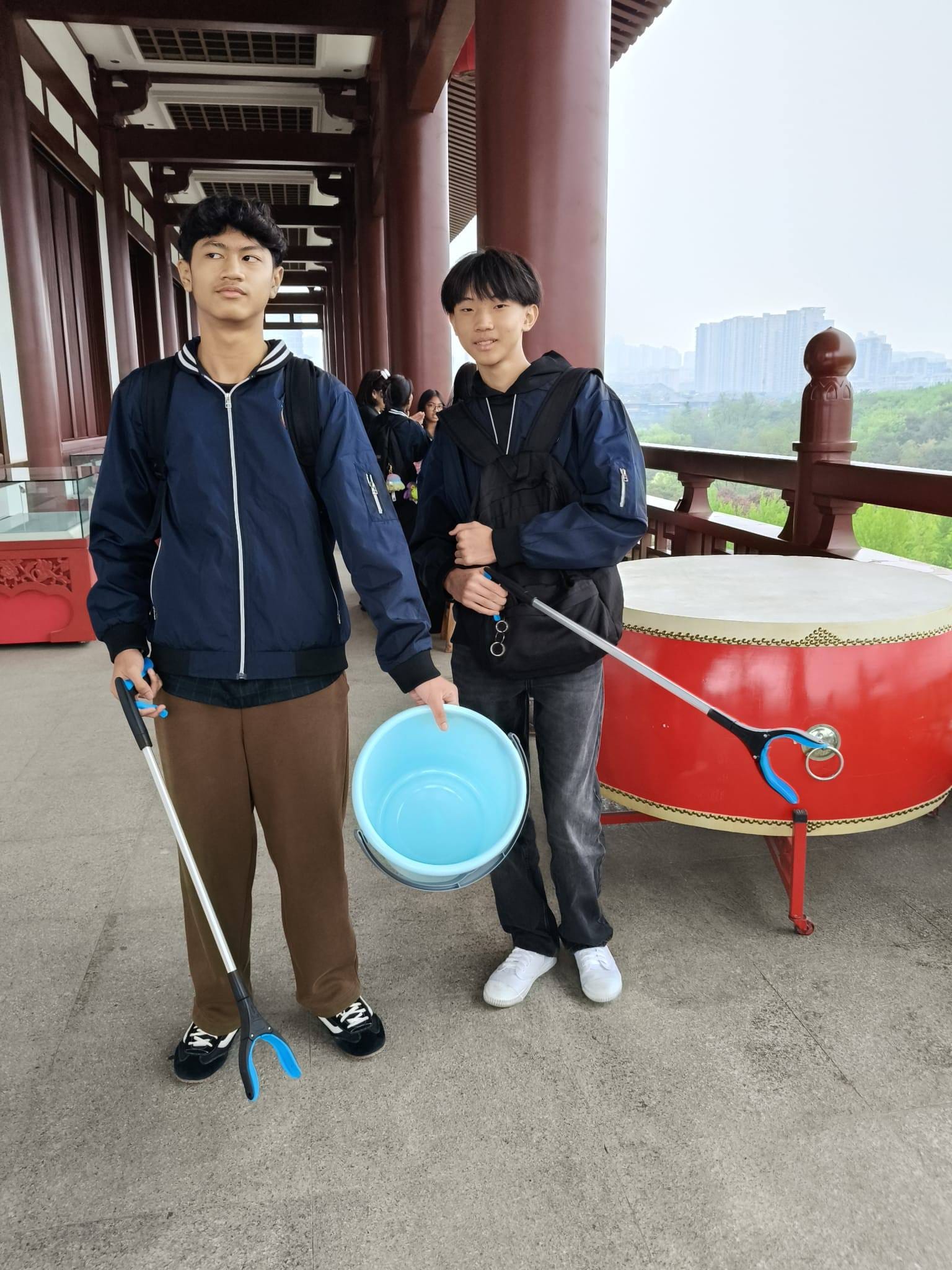 Two teenage boys in blue jackets hold a litter picker and blue bucket near a large red traditional Chinese drum.