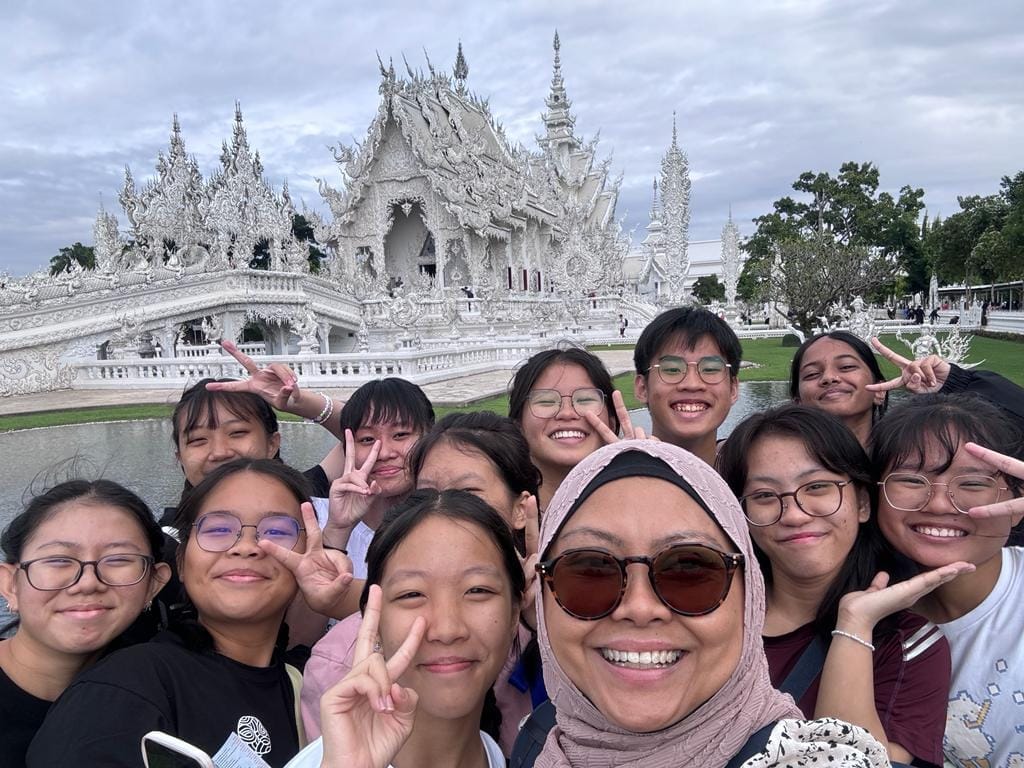 A group of smiling young people pose with peace signs in front of the ornate white Wat Rong Khun temple in Thailand.