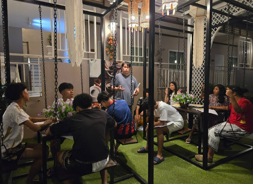 A group of people sit on swing seats at outdoor cafe tables while one person stands and speaks to them.