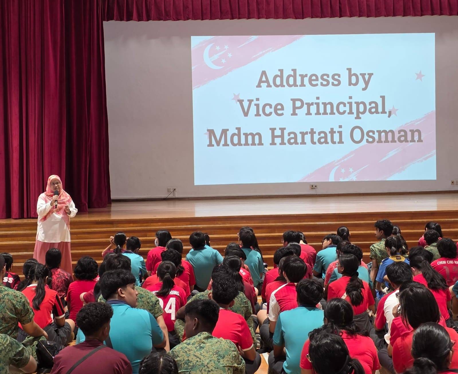 A woman in a pink hijab speaks to seated students in a hall; a screen reads "Address by Vice Principal, Mdm Hartati Osman."