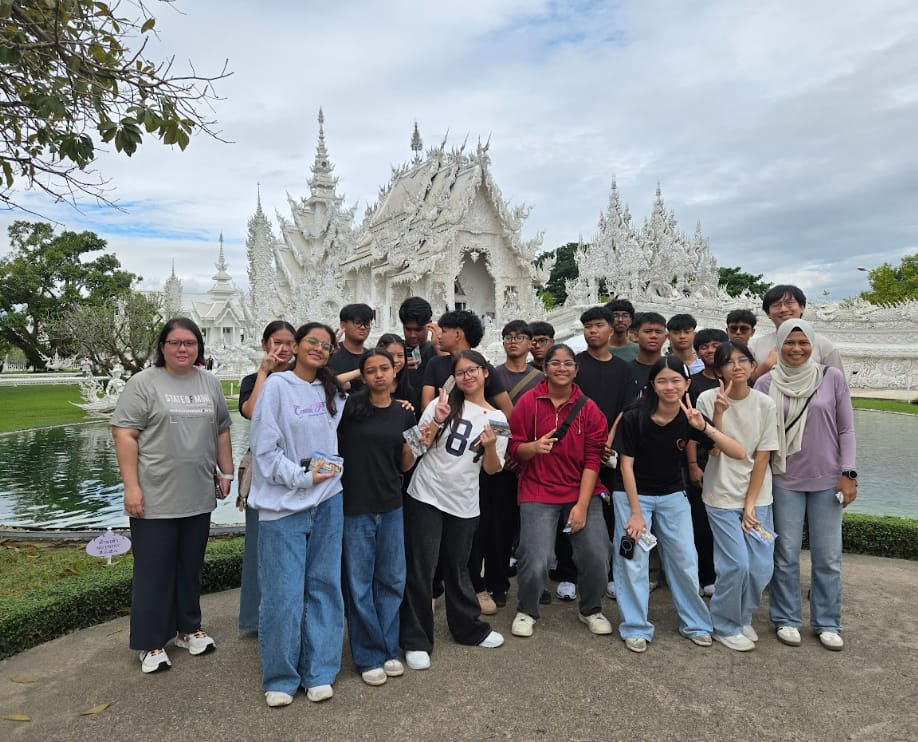 A group of about 20 young people posing in front of the ornate white Wat Rong Khun temple in Thailand.