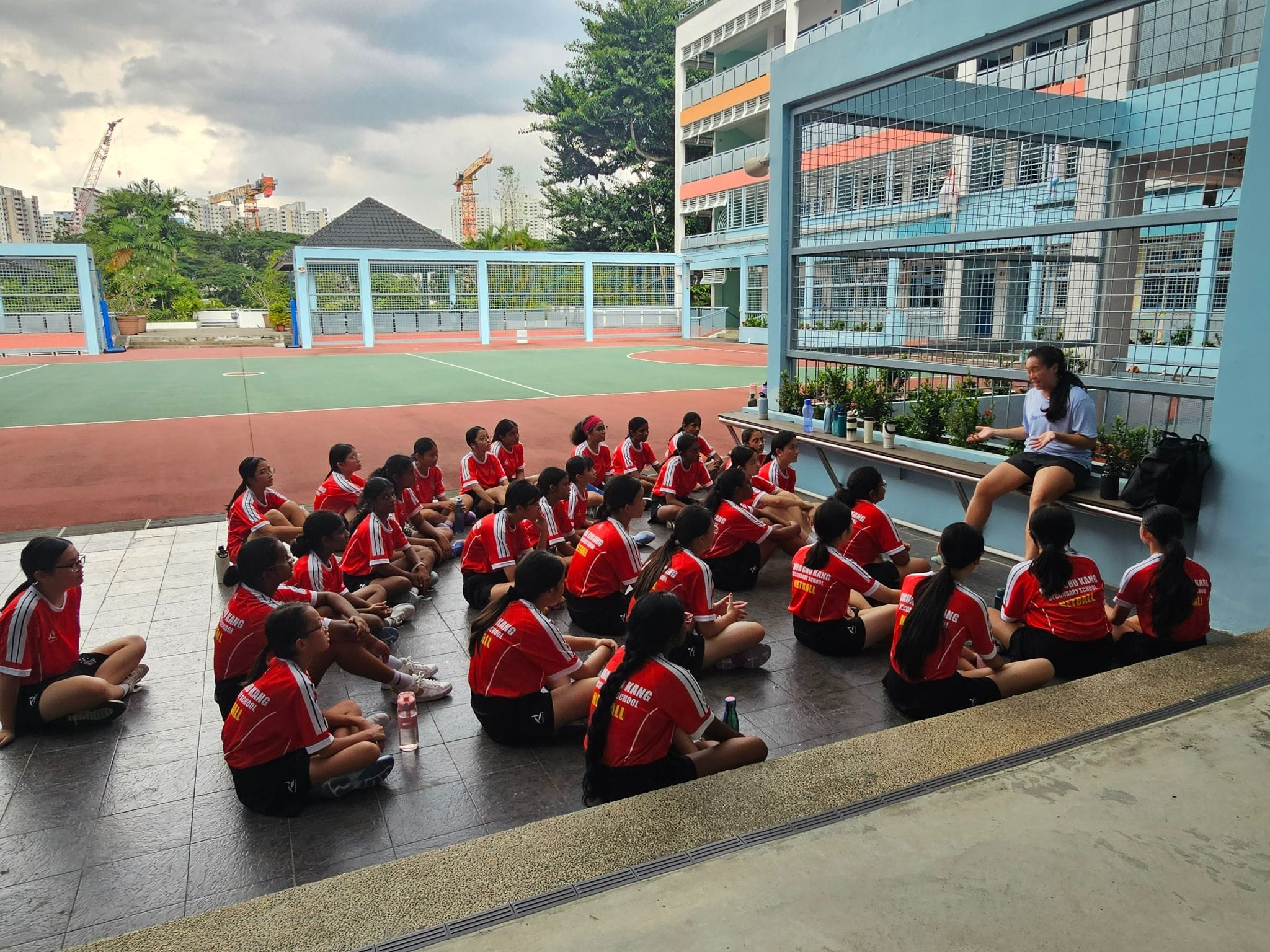 A group of girls in red school sports jerseys sit on the ground listening to a coach near an outdoor court.