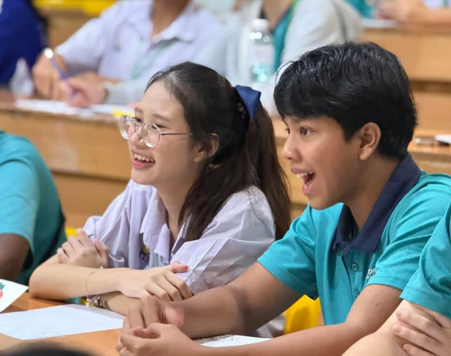 Two school students sit at a desk laughing and looking surprised in a busy classroom.