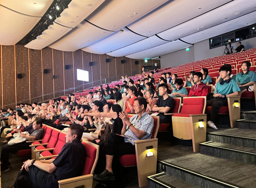 A crowd of people sitting in red and wood auditorium seats, many clapping, with a screen visible at the back.