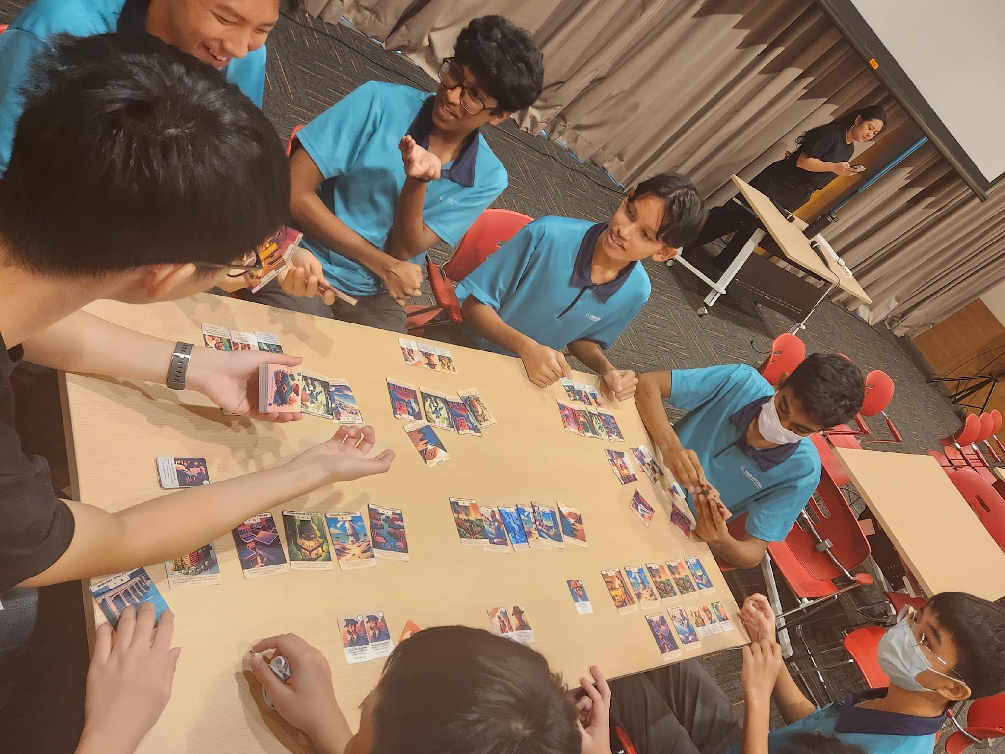 Several teens in blue shirts play a card game around a large table covered with colorful cards in a classroom.