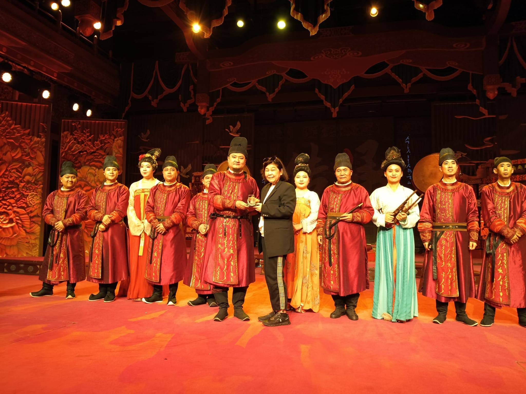 A group of performers in red traditional Chinese costumes stand on a red stage with ornate wooden backdrop.