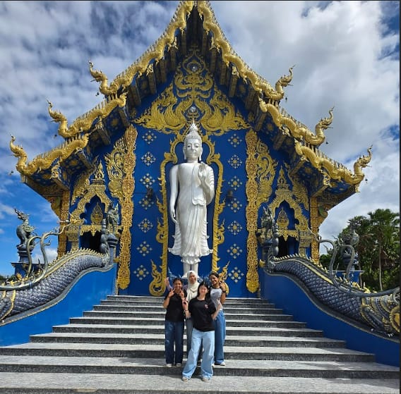 Three people stand on steps before a blue and gold Thai temple with a tall white Buddha statue.