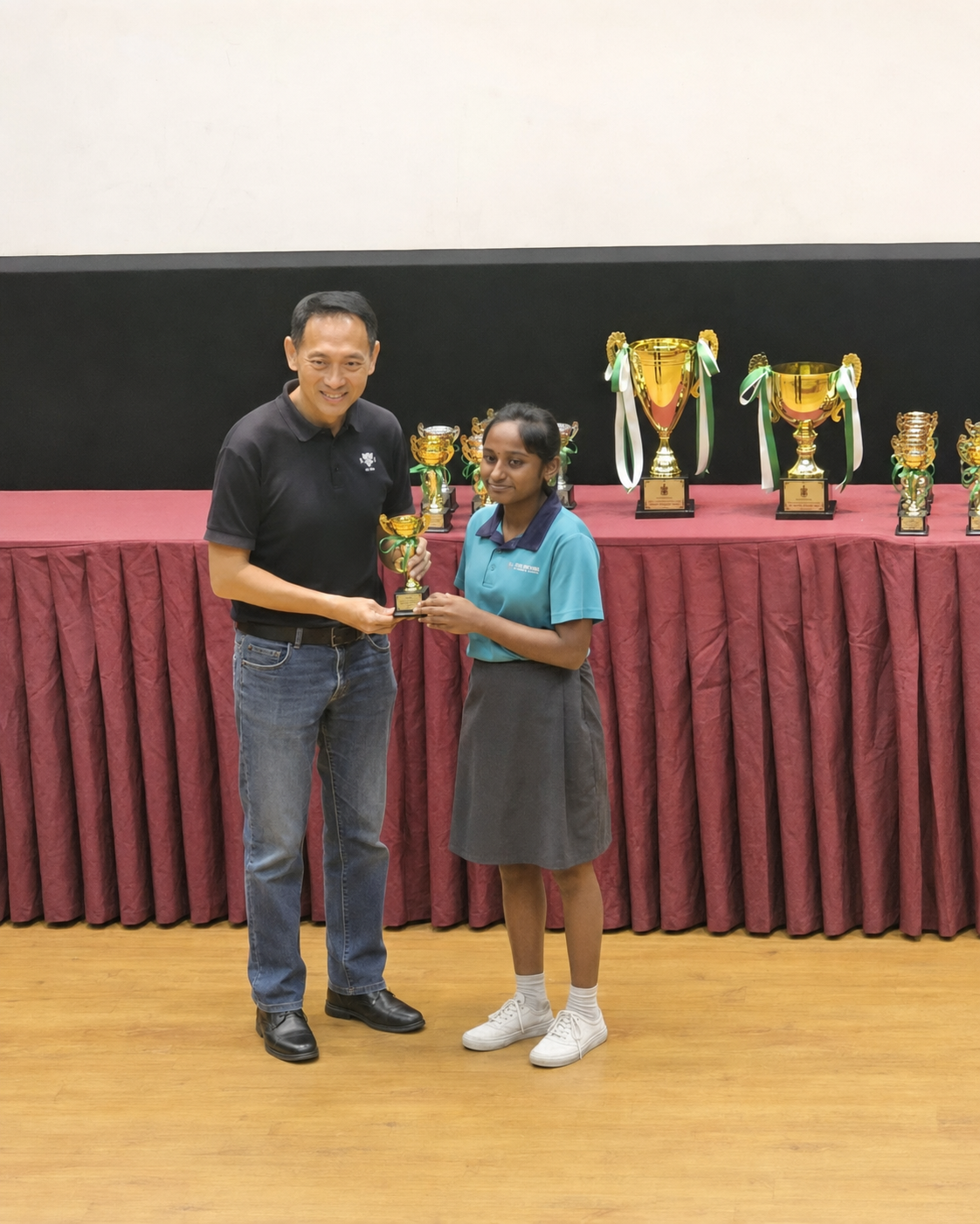 A man in a black polo shirt presents a gold trophy to a young girl in a teal shirt, with more trophies displayed behind them.