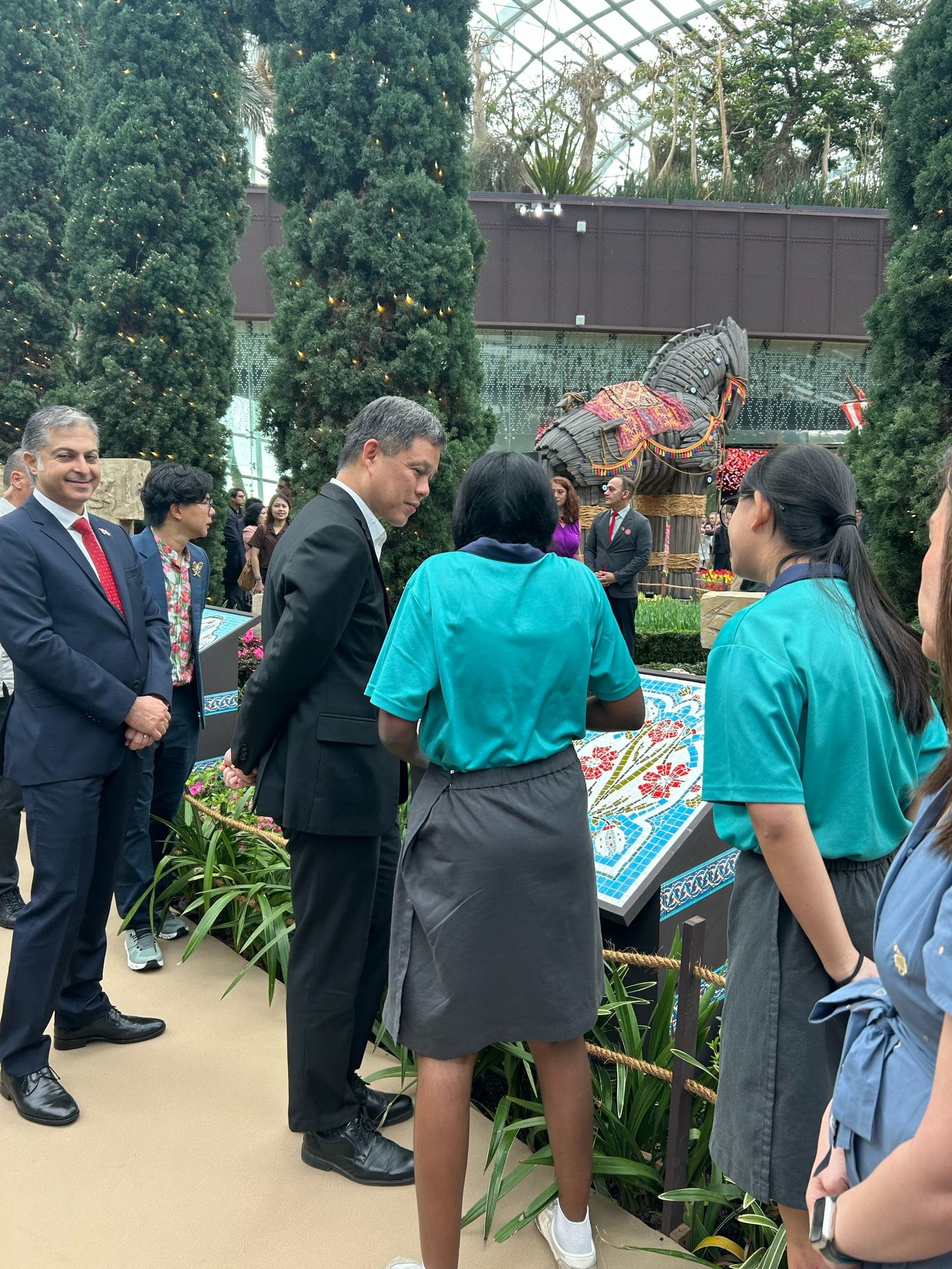 Adults in suits and students in teal uniforms gather indoors near floral displays and a large wooden horse sculpture.