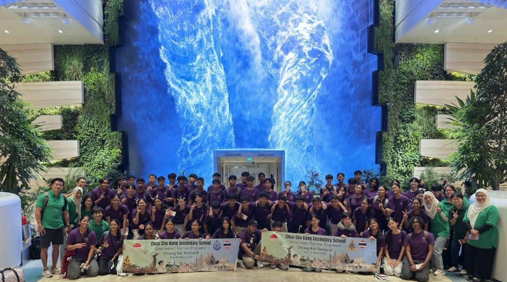 Large group of students in purple and green shirts pose before a tall blue waterfall display with green plant walls.