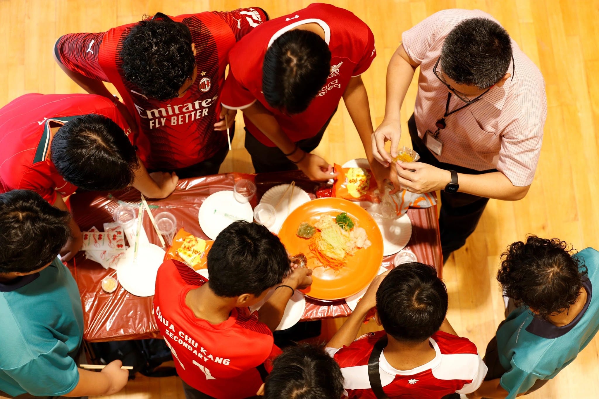 Overhead view of a group of youths and an adult in red shirts gathered around a table preparing yusheng salad.