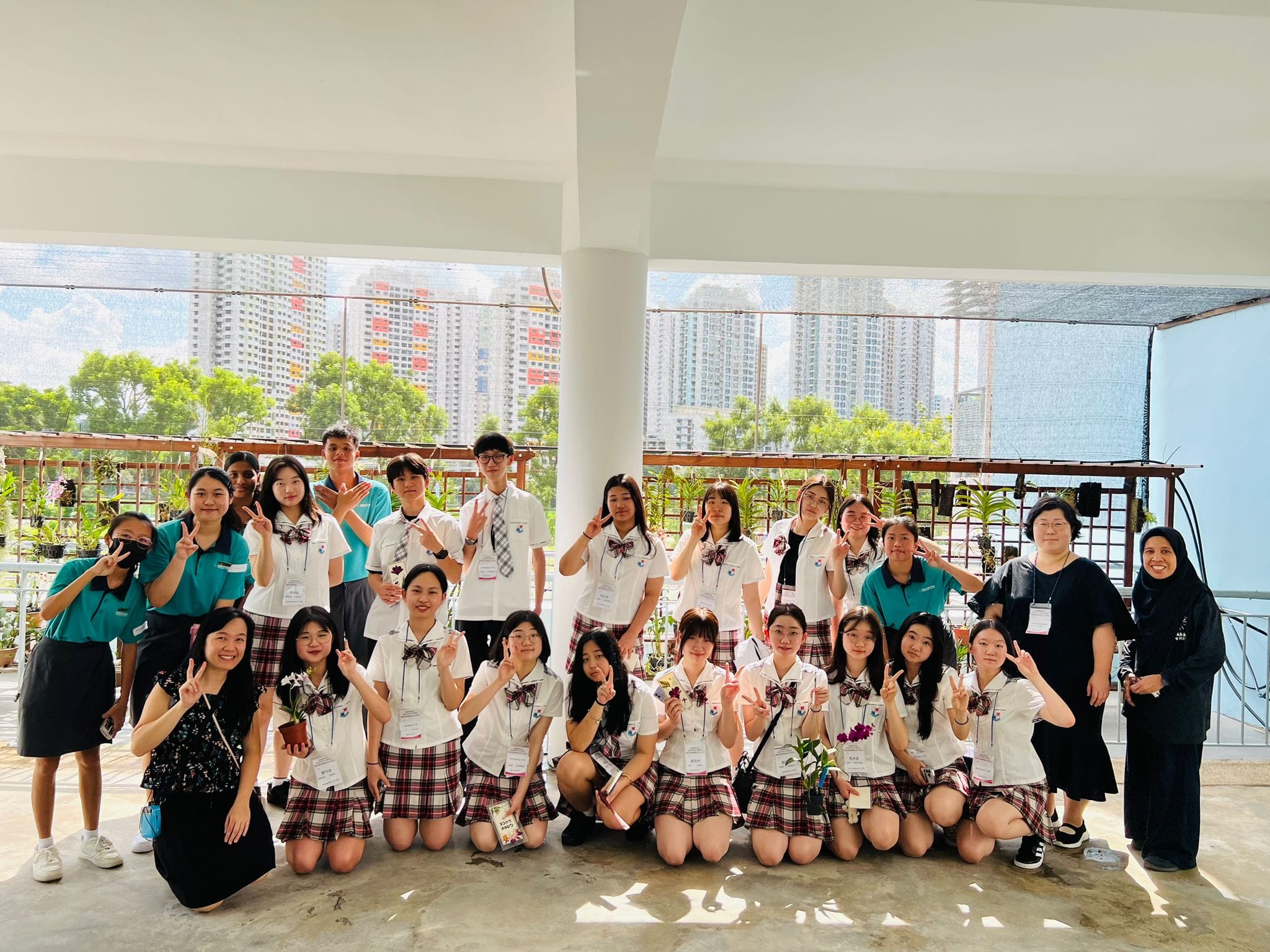 A large group of students in white and plaid uniforms pose with peace signs on an open terrace with city buildings behind.