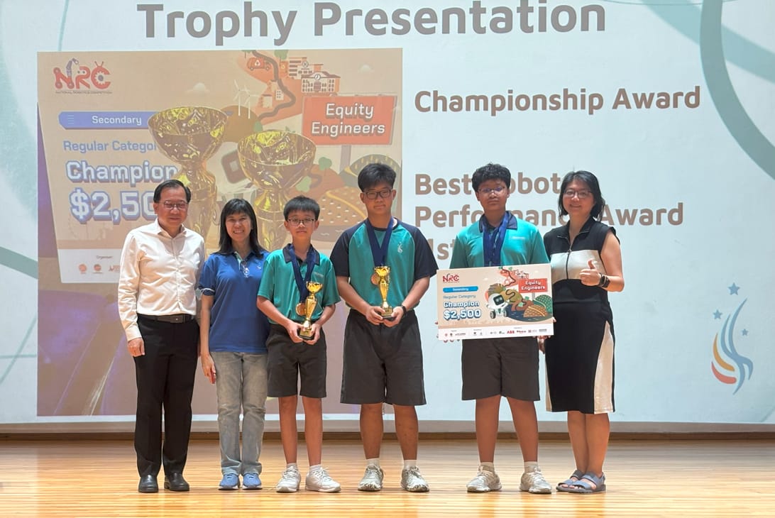 Six people stand on stage at an NRC Trophy Presentation, with students holding trophies and a $2,500 champion check.