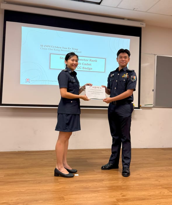 Two uniformed NPCC cadets stand together holding a certificate in front of a presentation screen.