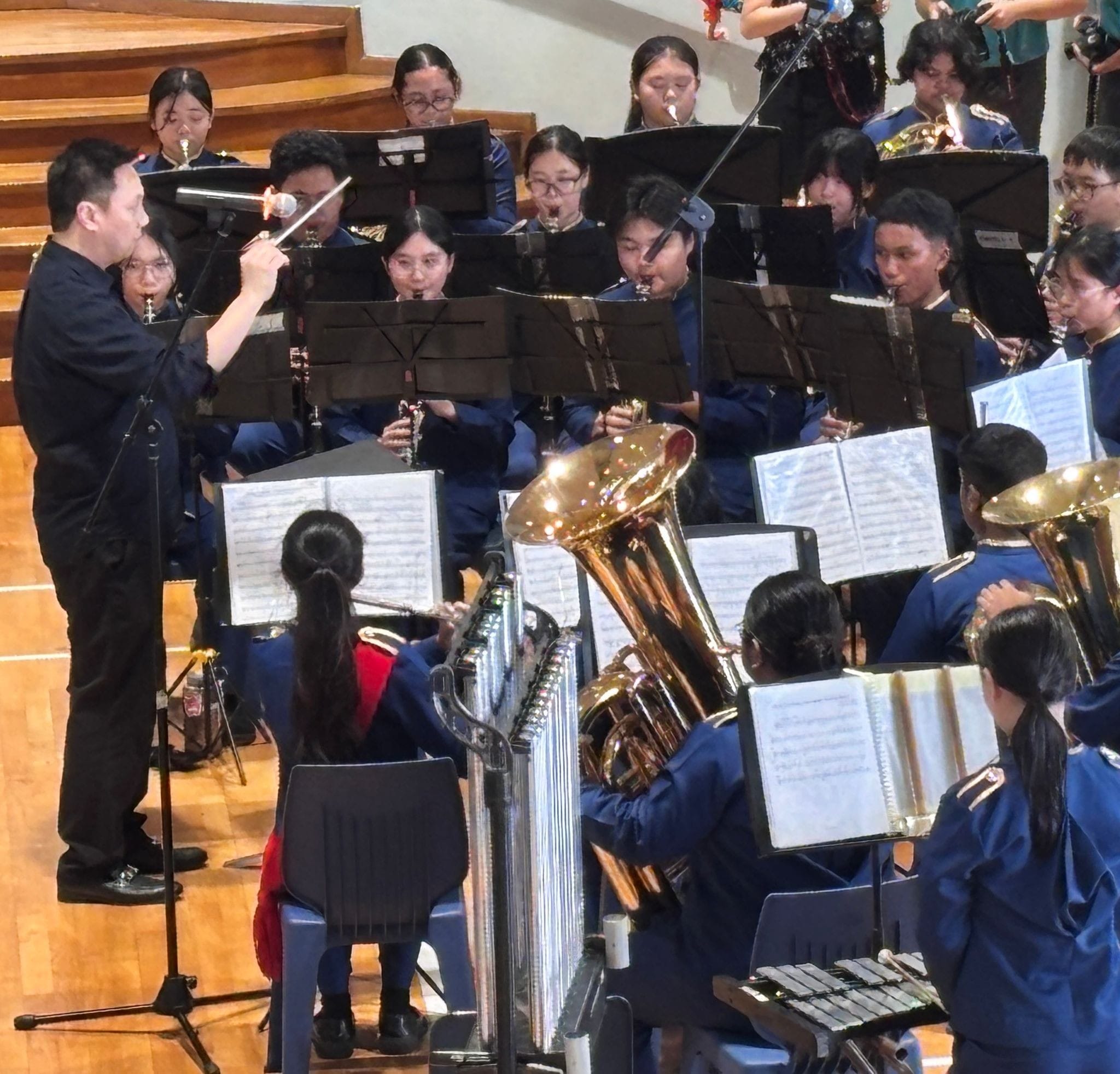 A conductor in black leads a youth wind band in blue uniforms playing brass and woodwind instruments on stage.