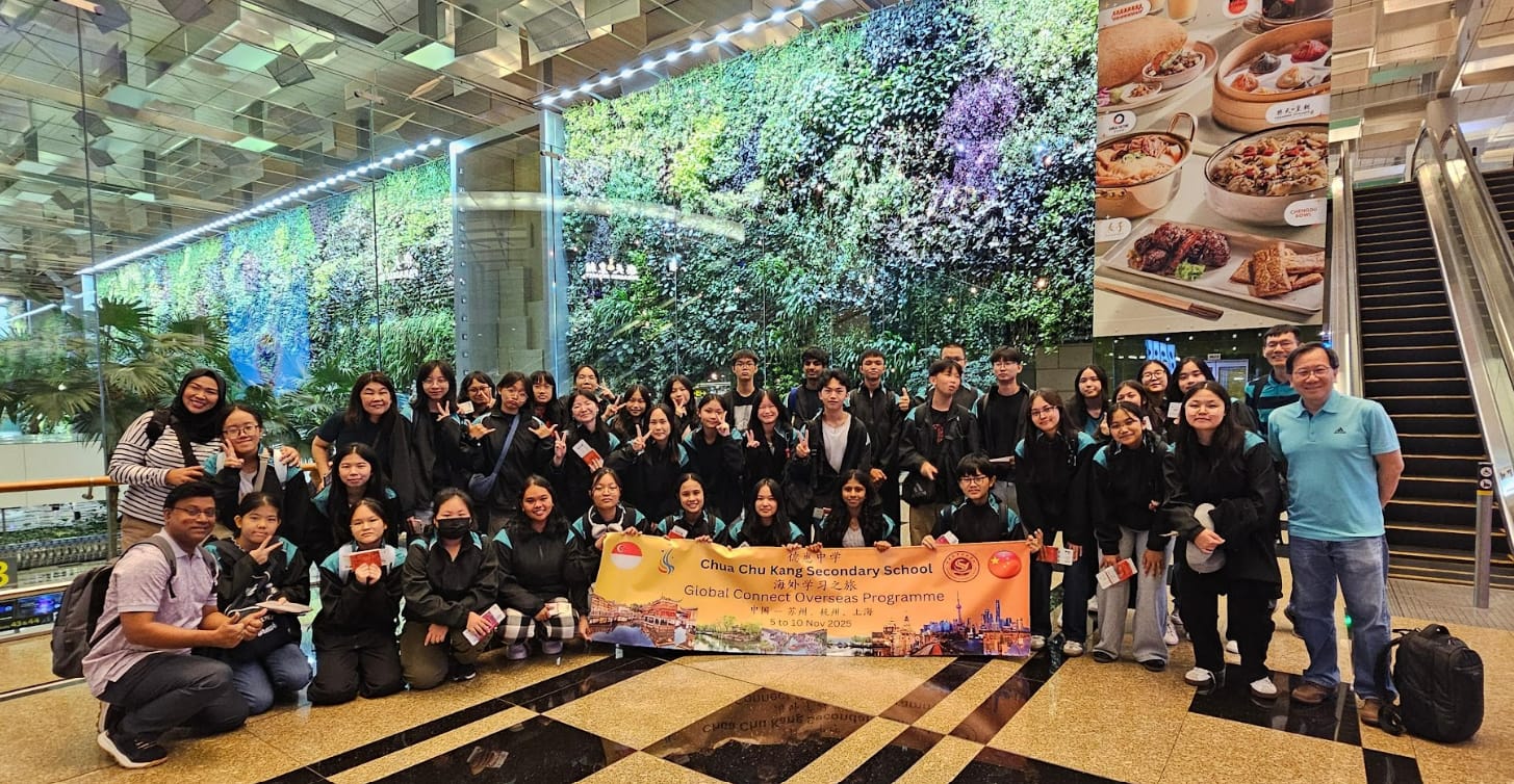Large student group poses with a Chua Chu Kang Secondary School overseas programme banner in front of a green living wall.