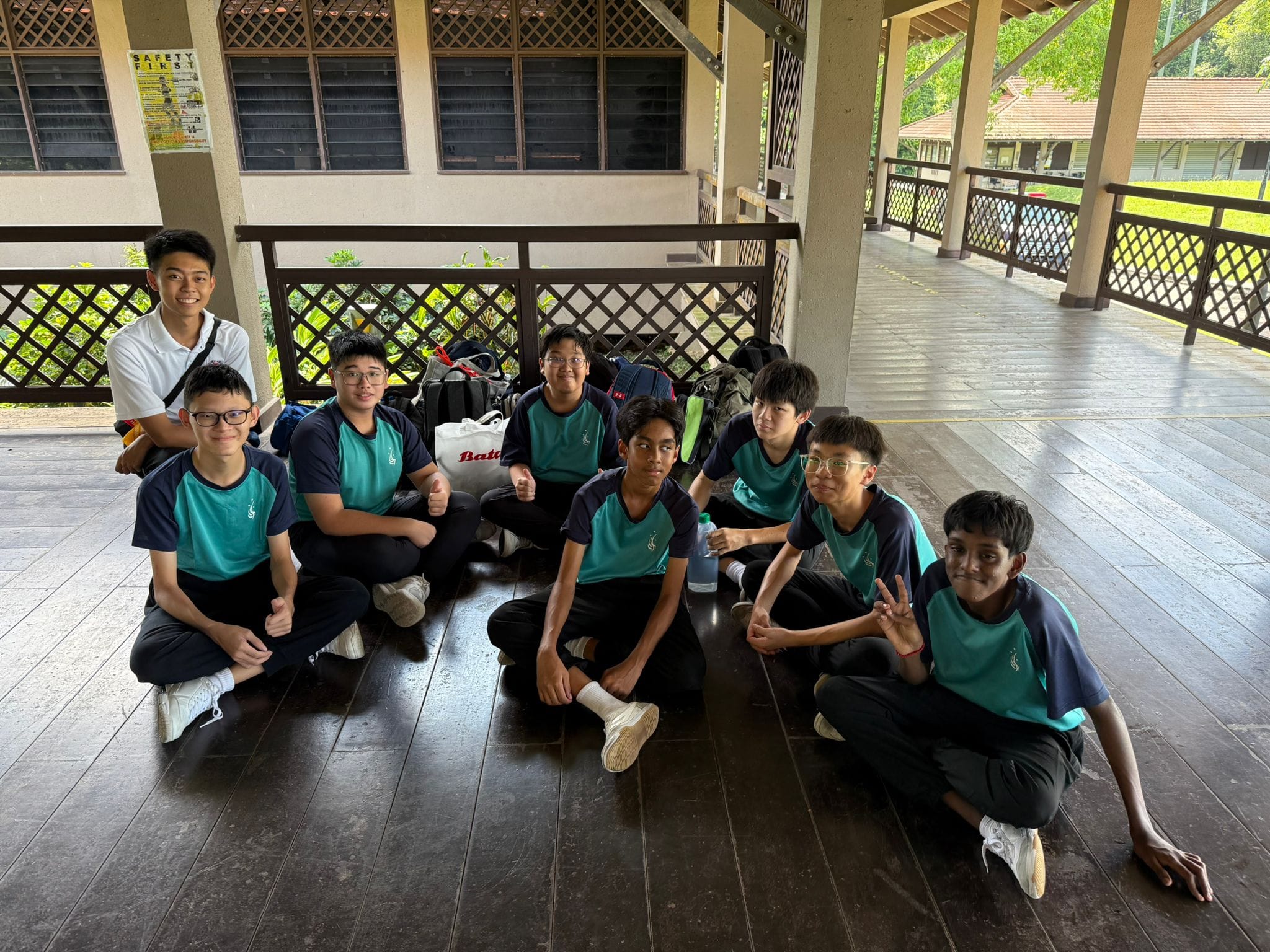 Eight boys in matching teal and navy uniforms sit on a wooden floor of an outdoor covered pavilion, smiling at the camera.